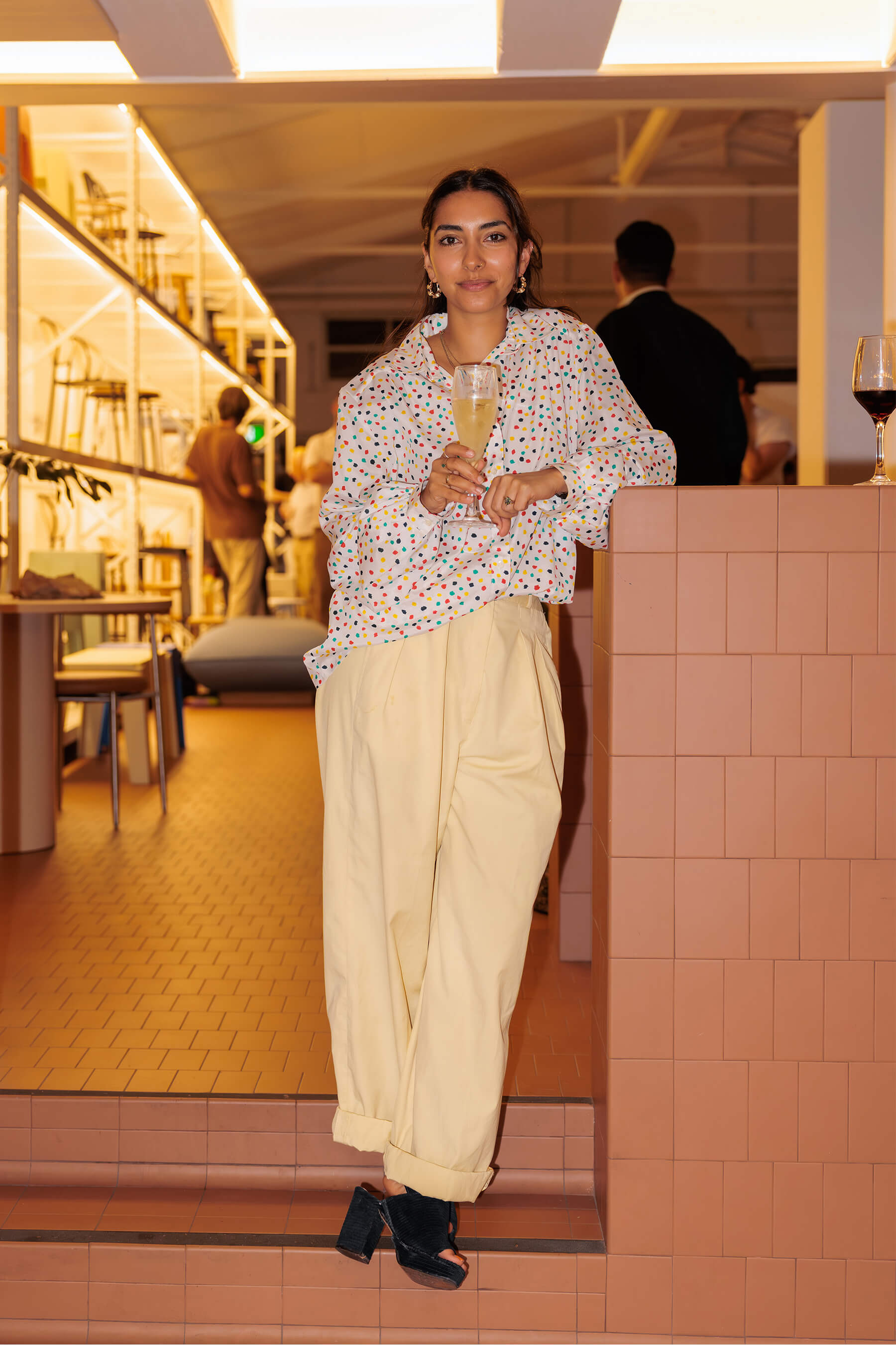 A guest poses with a champagne glass beside the tiled bar at the DesignByThem “I’ll Be Happy When” event, surrounded by chairs and shelving displays.