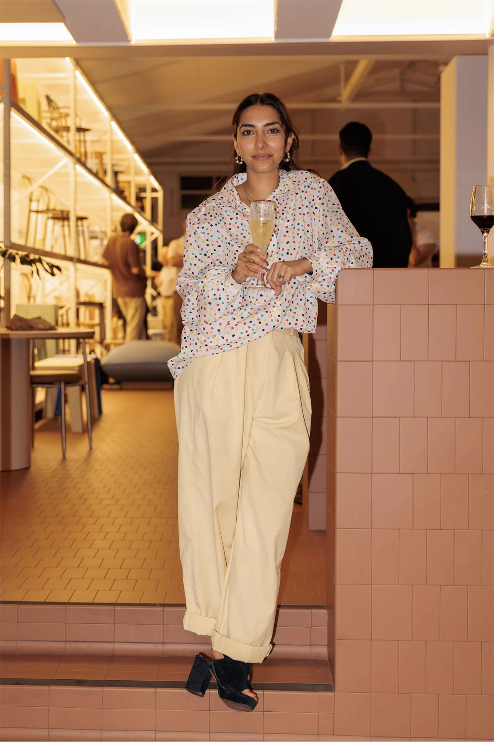 A guest poses with a champagne glass beside the tiled bar at the DesignByThem “I’ll Be Happy When” event, surrounded by chairs and shelving displays.