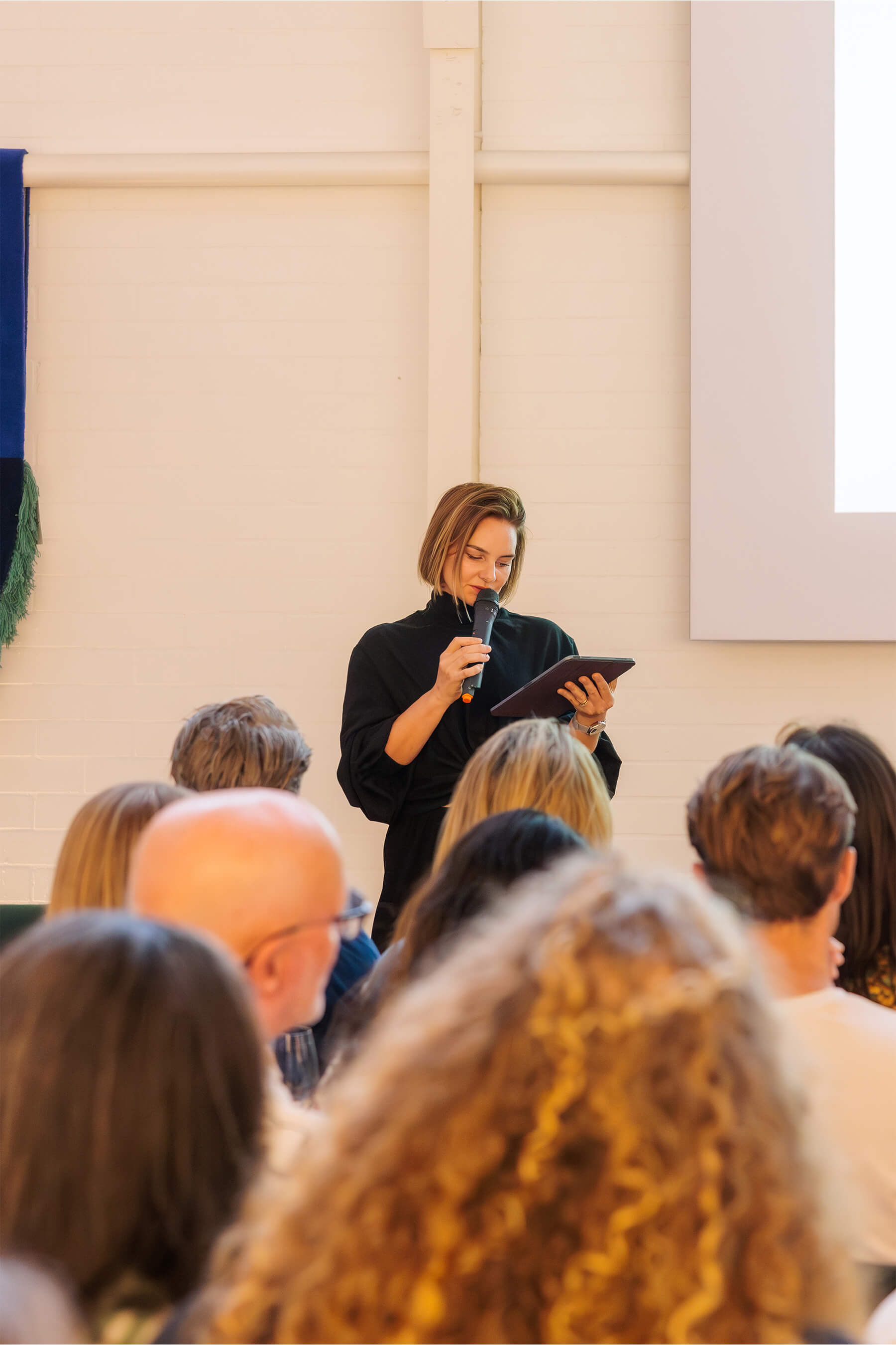 Sarah Gibson reads from a tablet while speaking into a microphone, standing in front of a seated crowd during the event.