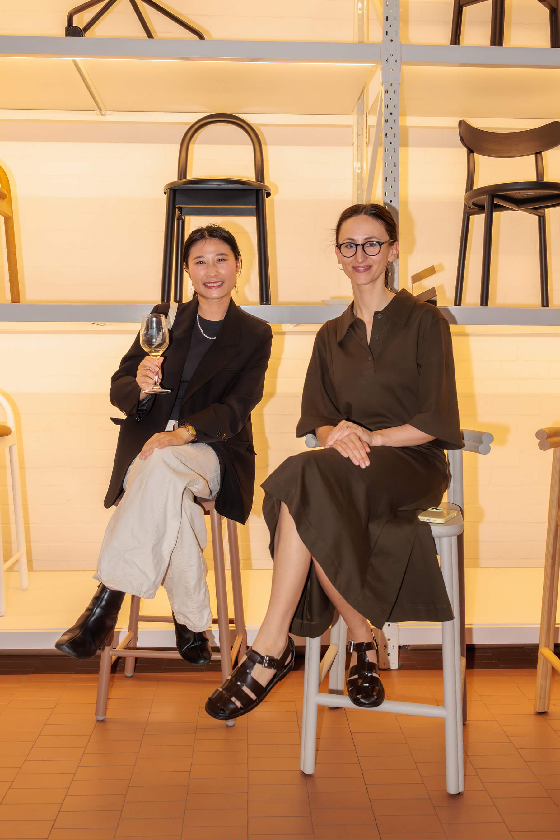 Two attendees seated in front of a shelving wall filled with designer chairs, smiling and holding drinks at the DesignByThem studio.