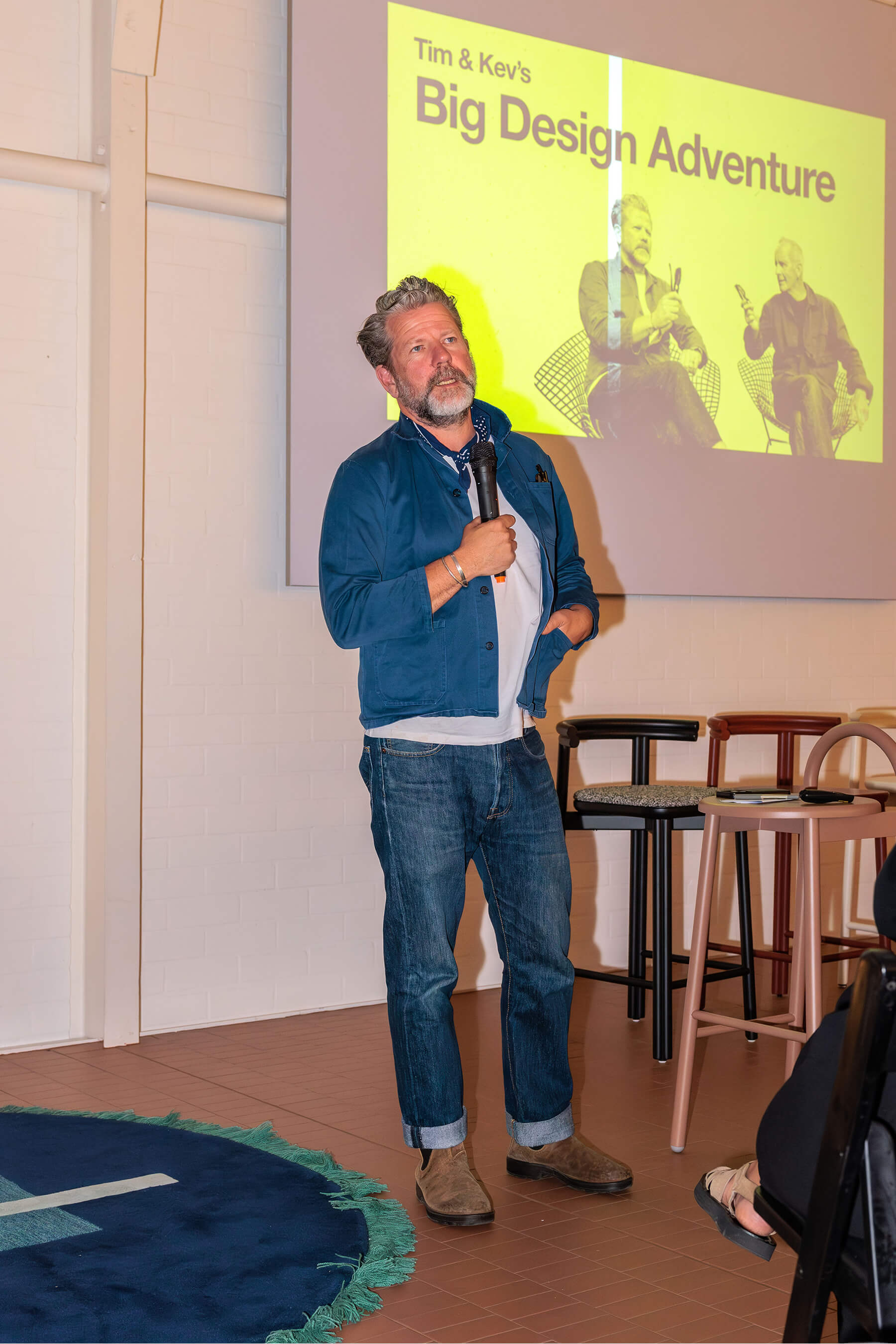Tim Ross stands on stage holding a microphone during his talk, with the projected slide behind him reading “Tim & Kev’s Big Design Adventure”.