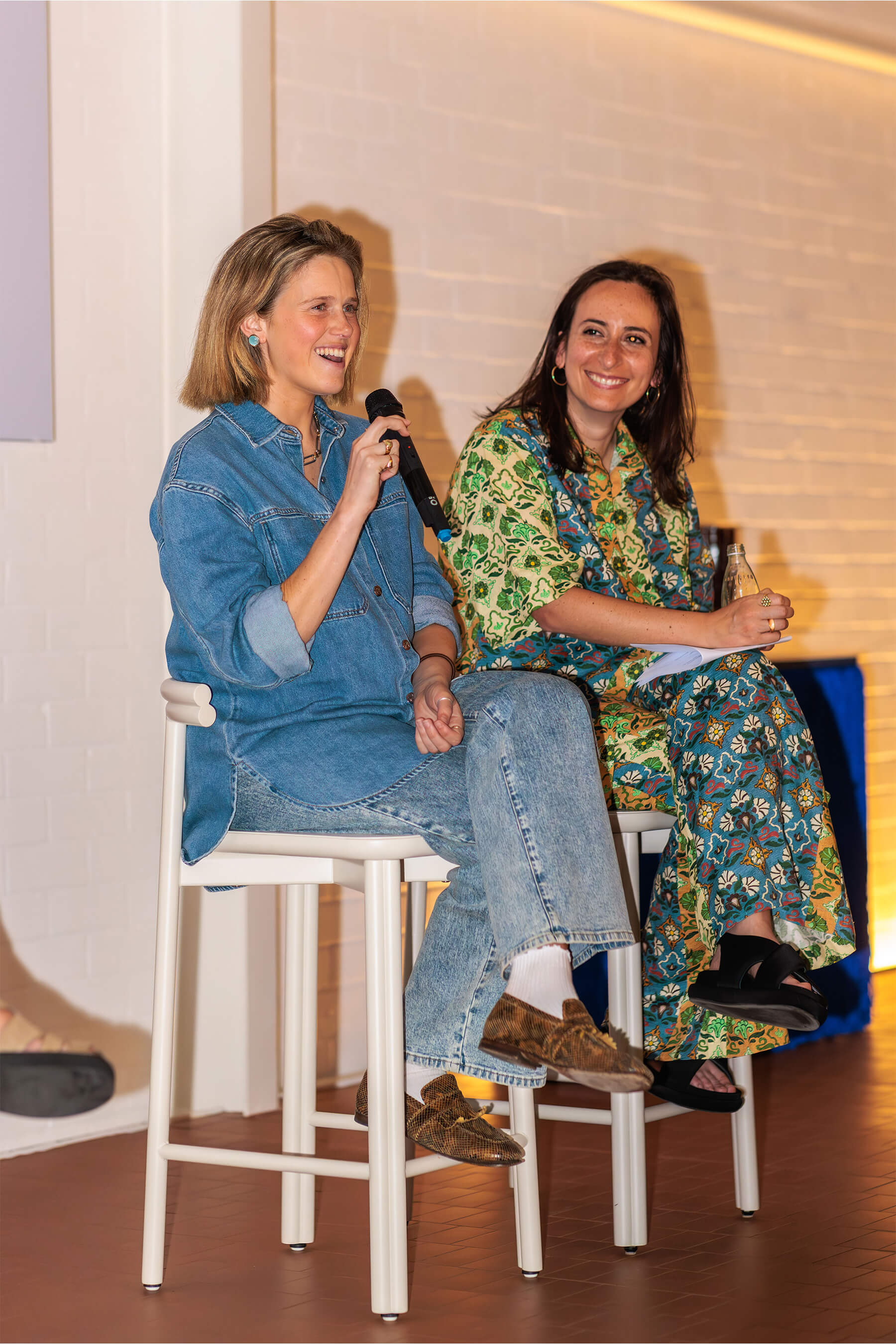 Sarah Gibson and Eliza Gosse smiling and speaking into microphones on stage, seated on high stools against a softly lit brick wall.