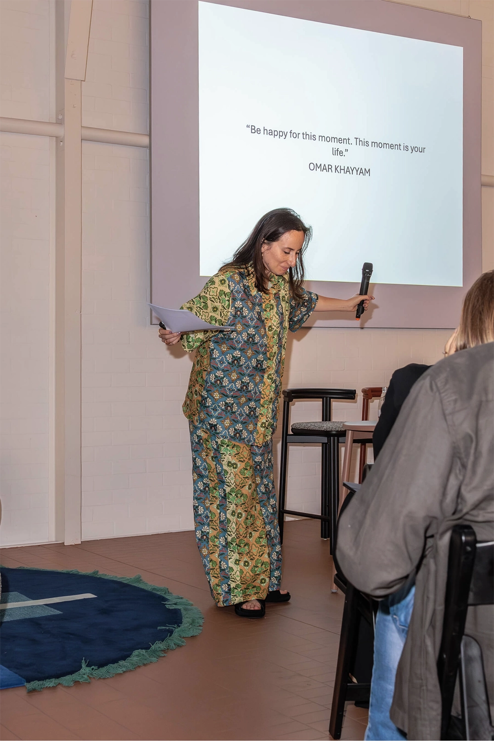 Nasim Koerting on stage holding a microphone and a sheet of notes beneath a projected quote: “Be happy for this moment. This moment is your life.”