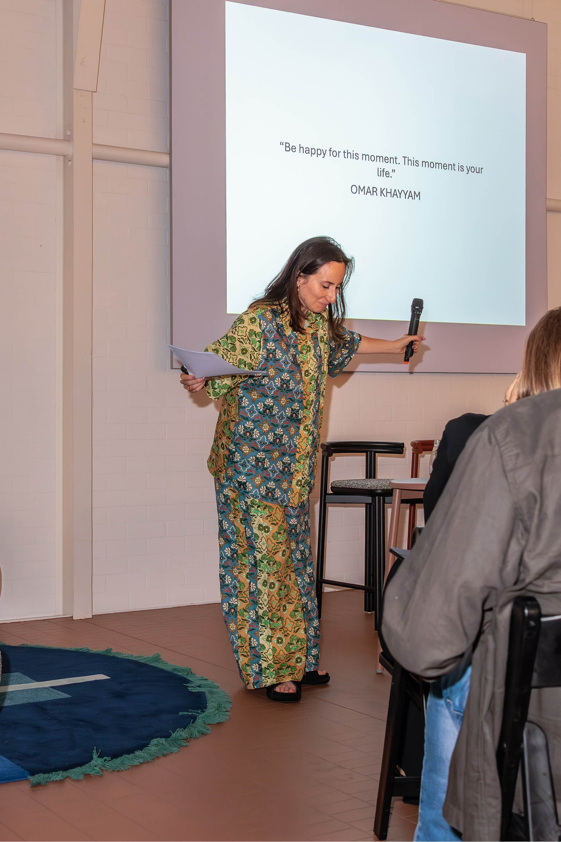 Nasim Koerting on stage holding a microphone and a sheet of notes beneath a projected quote: “Be happy for this moment. This moment is your life.”