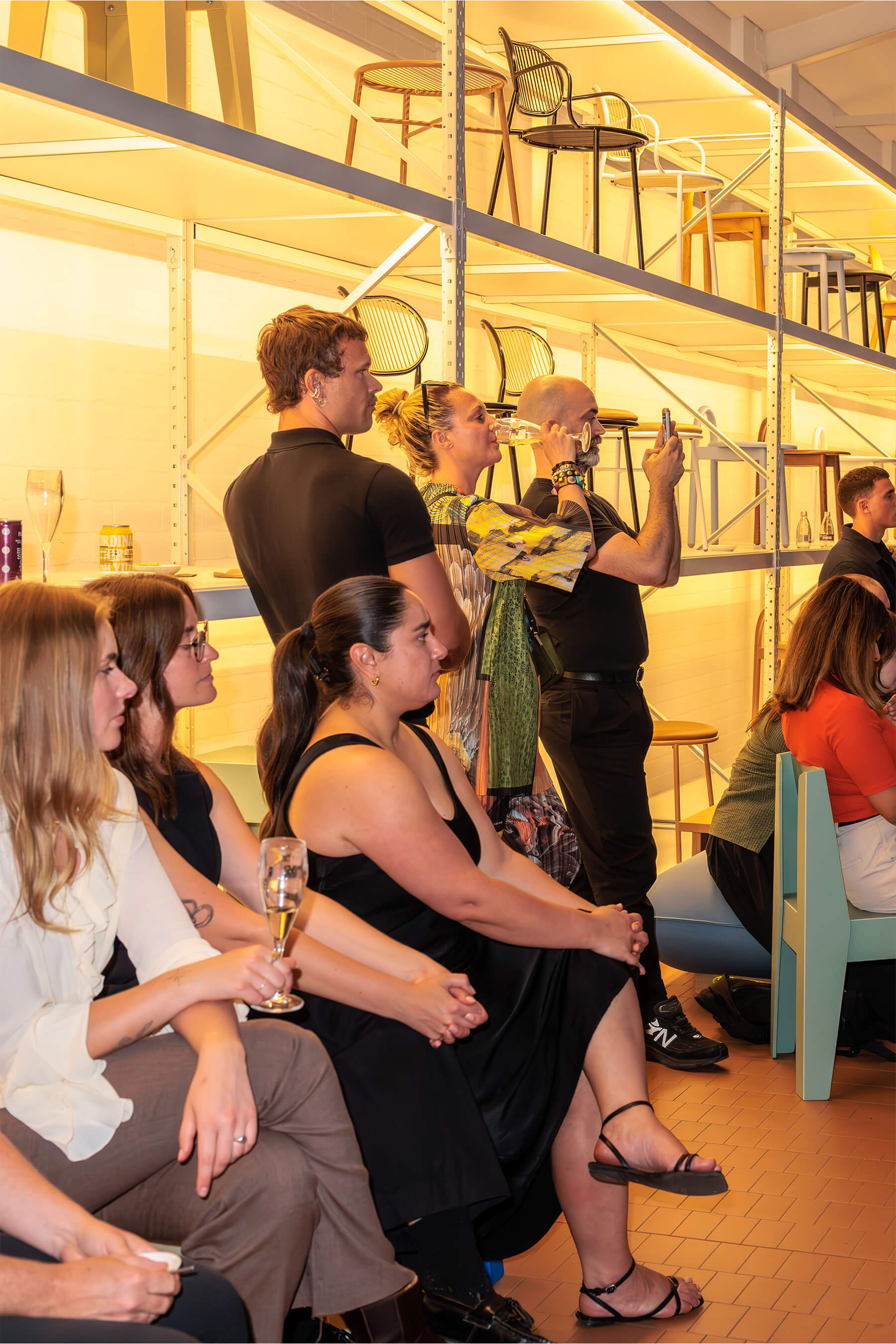 Audience members seated beside a backlit shelving wall filled with designer chairs, some drinking and taking photos during the panel.