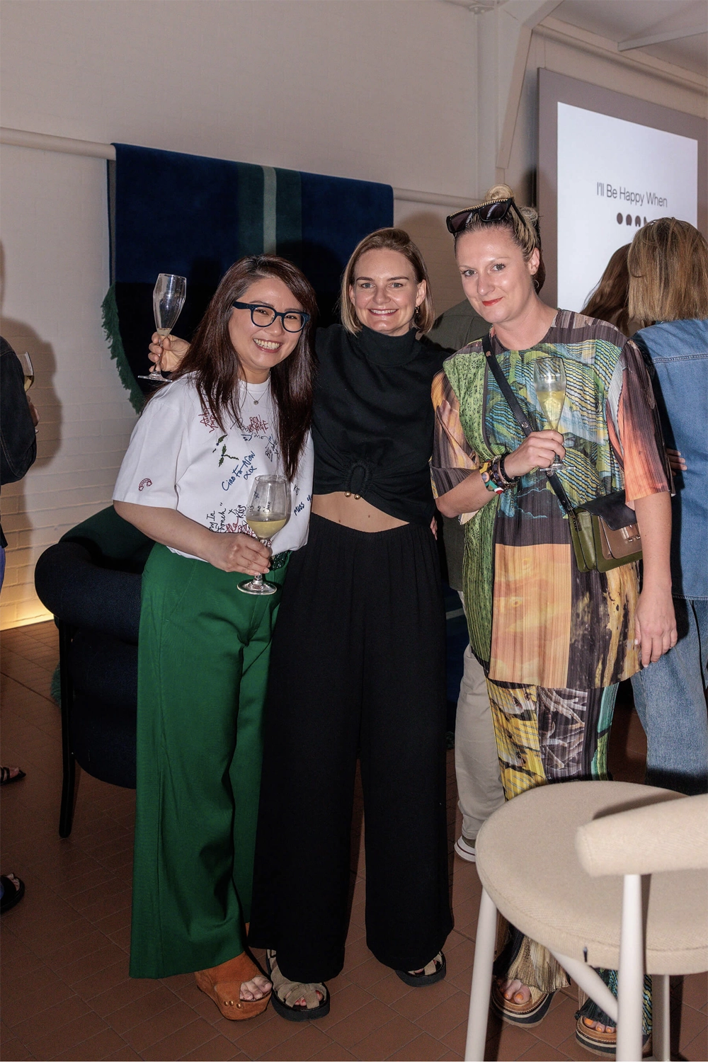 Three women smiling and holding drinks at the DesignByThem “I’ll Be Happy When” event, with a projected title screen in the background.