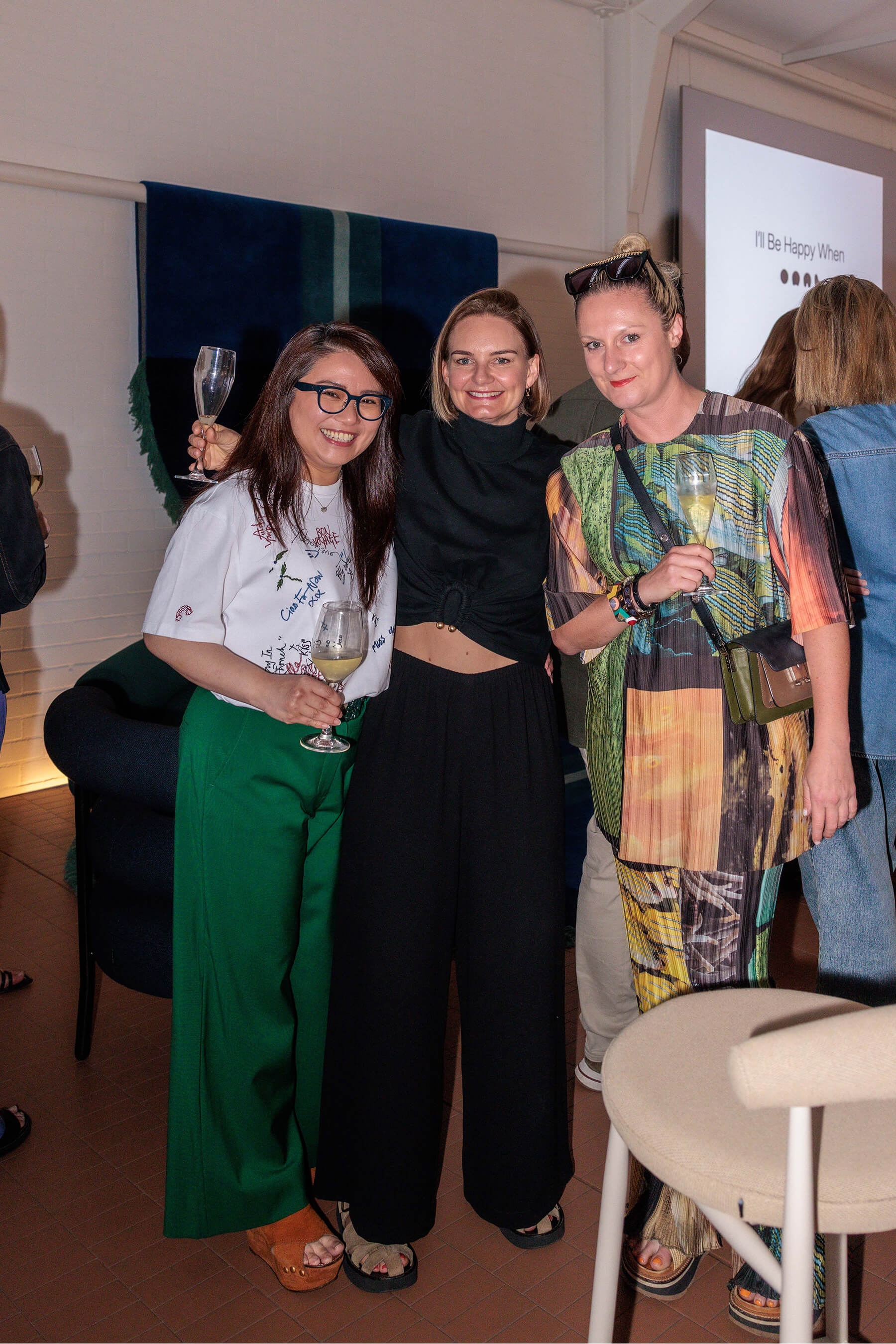 Three women smiling and holding drinks at the DesignByThem “I’ll Be Happy When” event, with a projected title screen in the background.