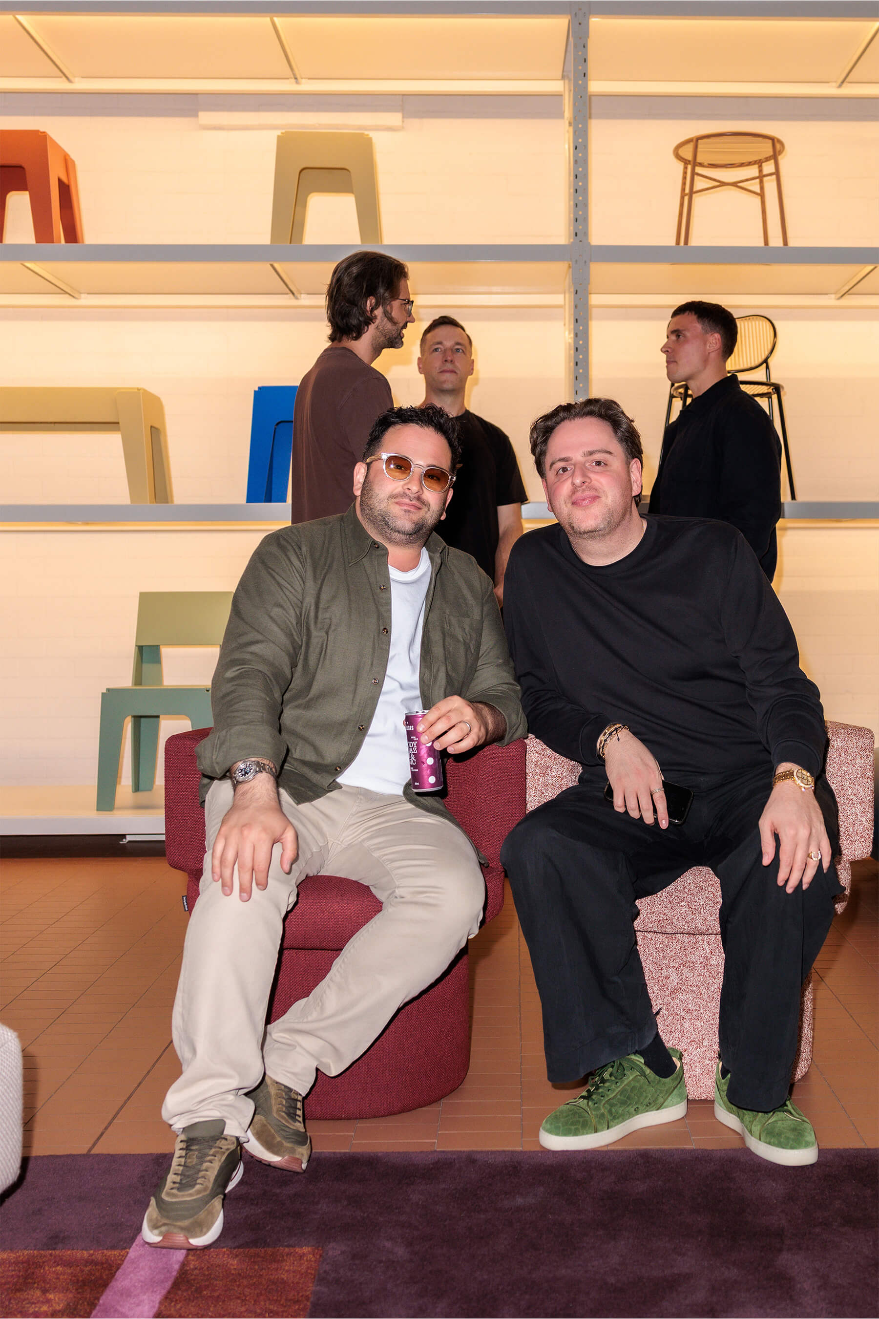 Two guests seated on contemporary furniture at the DesignByThem event, with others interacting in front of a backlit wall of colourful chairs and stools.