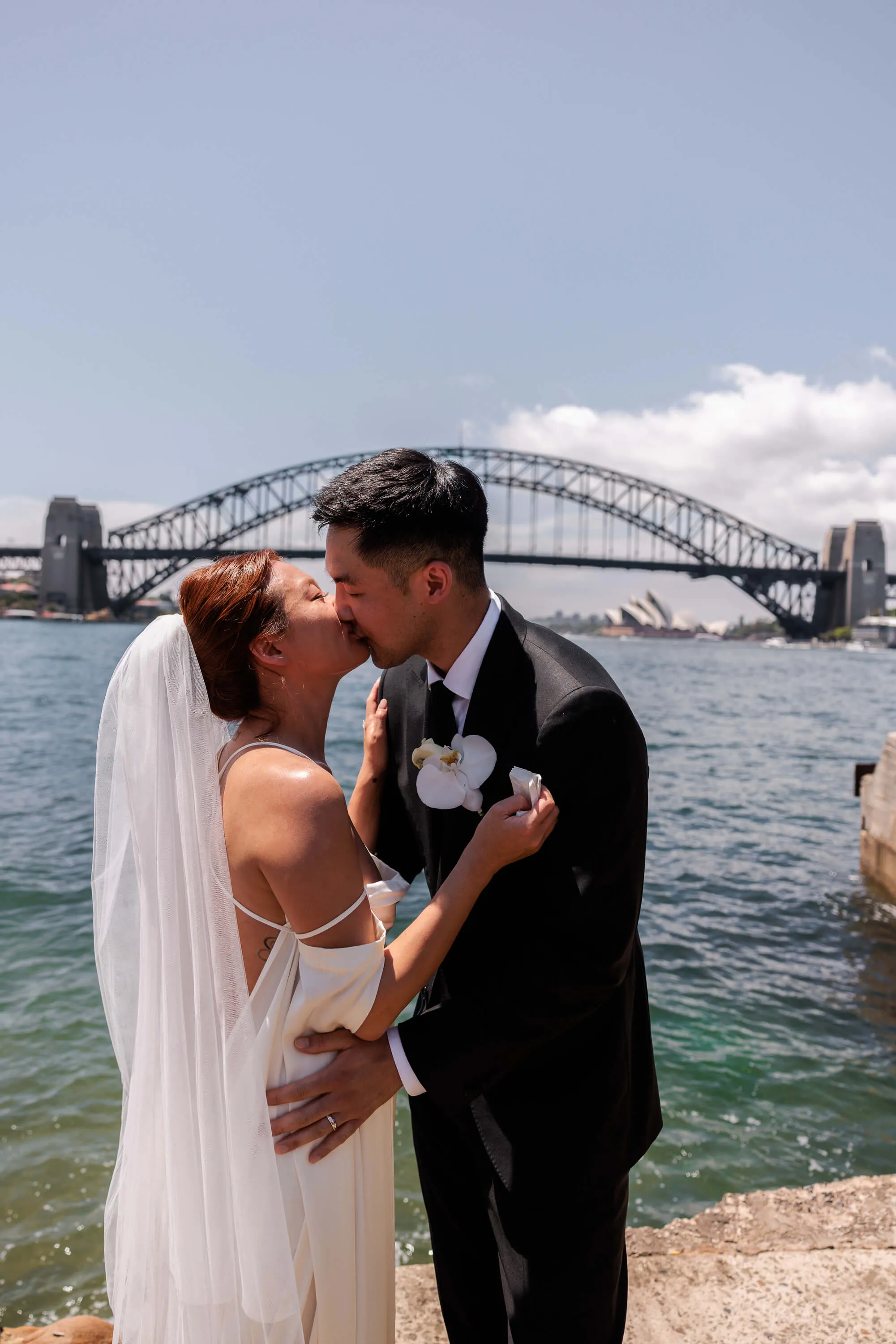 Melanie and Dai share their first kiss as newlyweds, standing by the water beneath the Sydney Harbour Bridge | EDG Studios