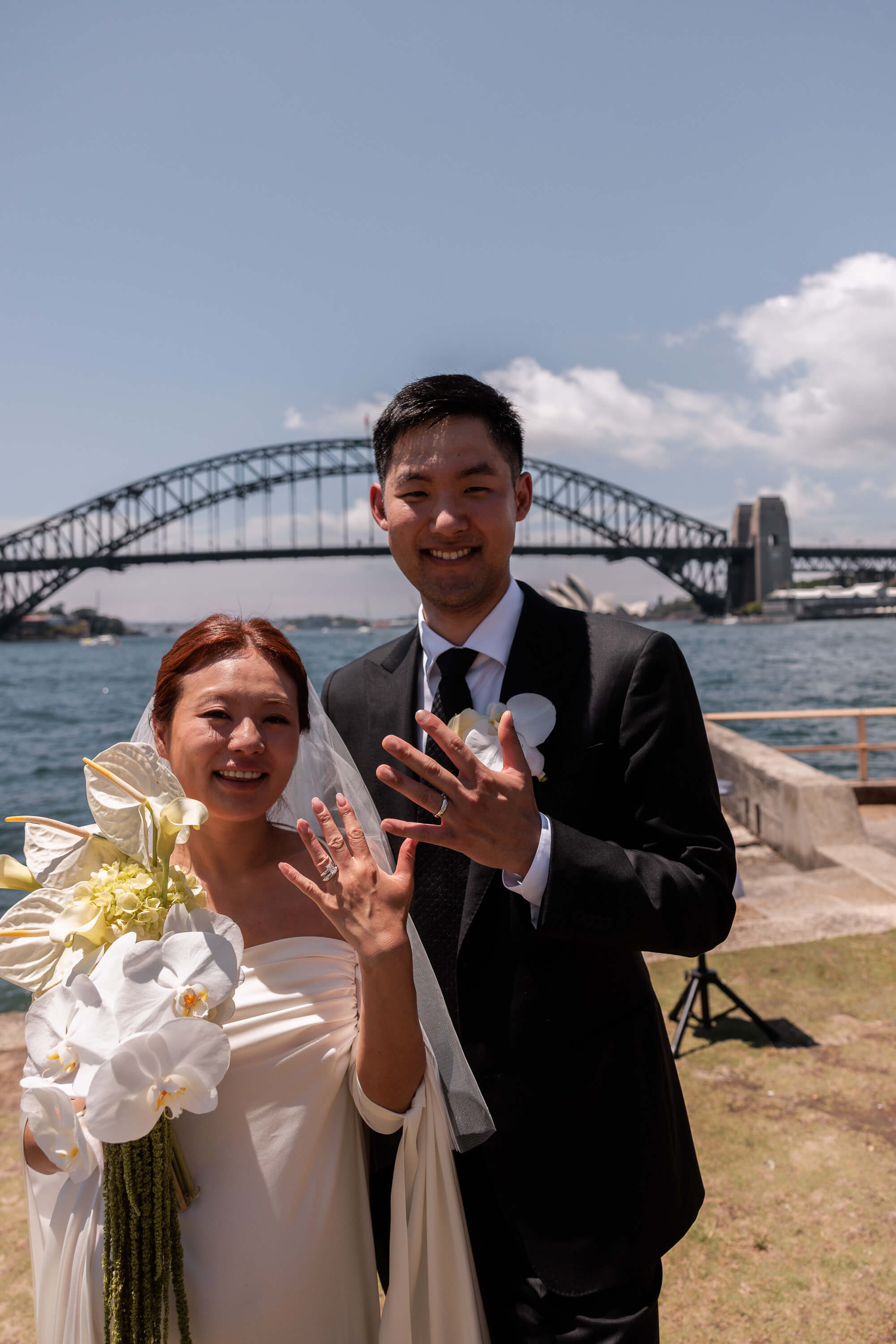 Melanie and Dai stand together holding their hands up to display their wedding rings, framed by Sydney Harbour Bridge.