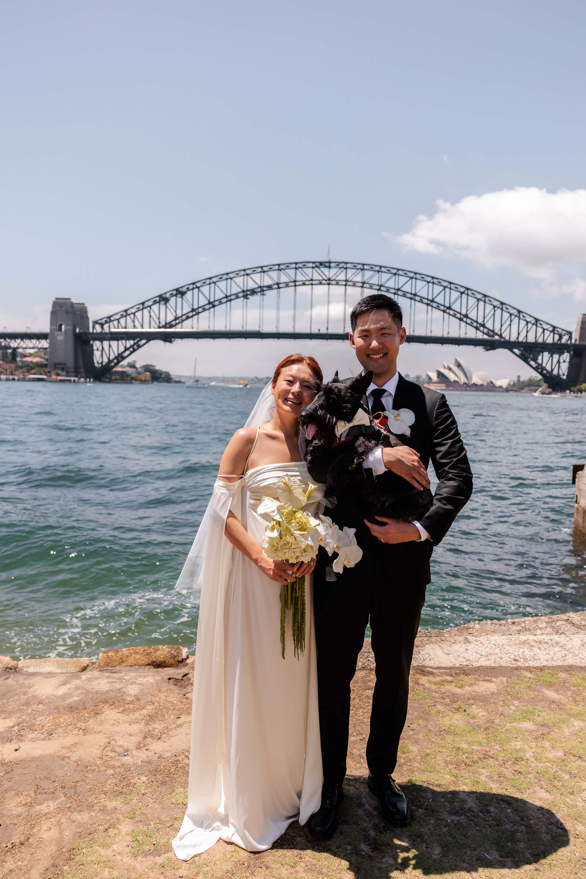 Melanie and Dai stand together with their dog following their wedding ceremony, with Sydney Harbour Bridge and the Opera House behind them.