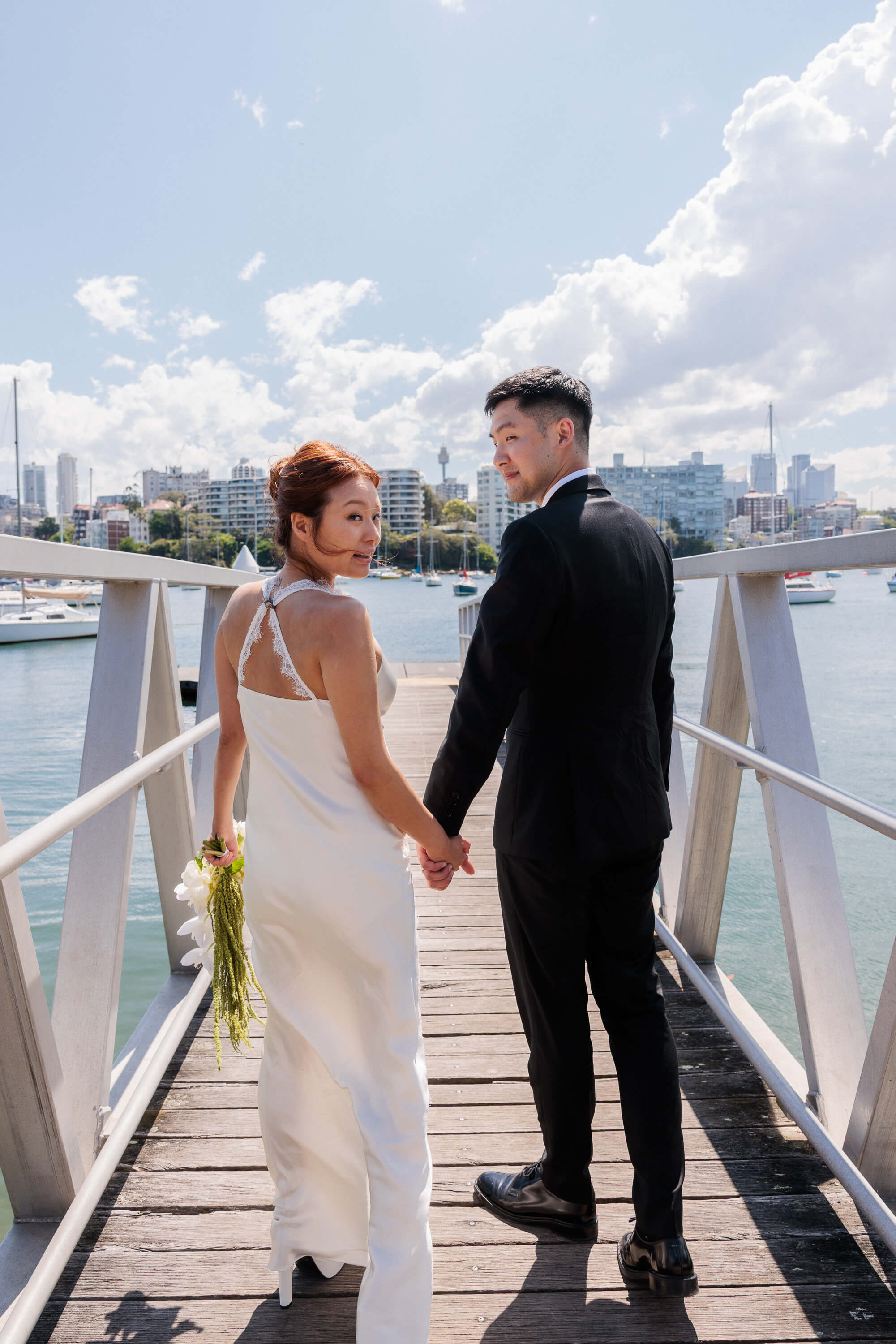 Melanie and Dai walk hand in hand along a marina gangway following their wedding ceremony, city skyline in the distance.