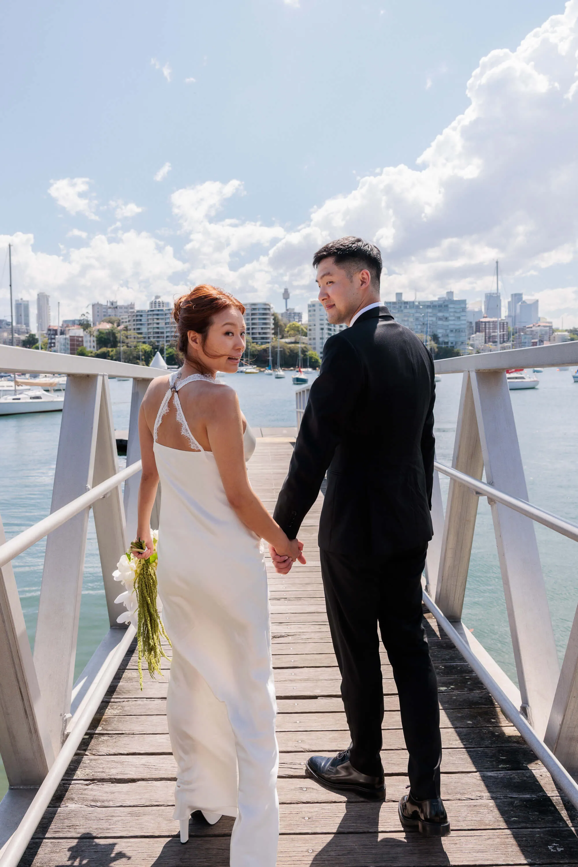 Melanie and Dai walk hand in hand along a marina gangway following their wedding ceremony, city skyline in the distance | EDG Studios