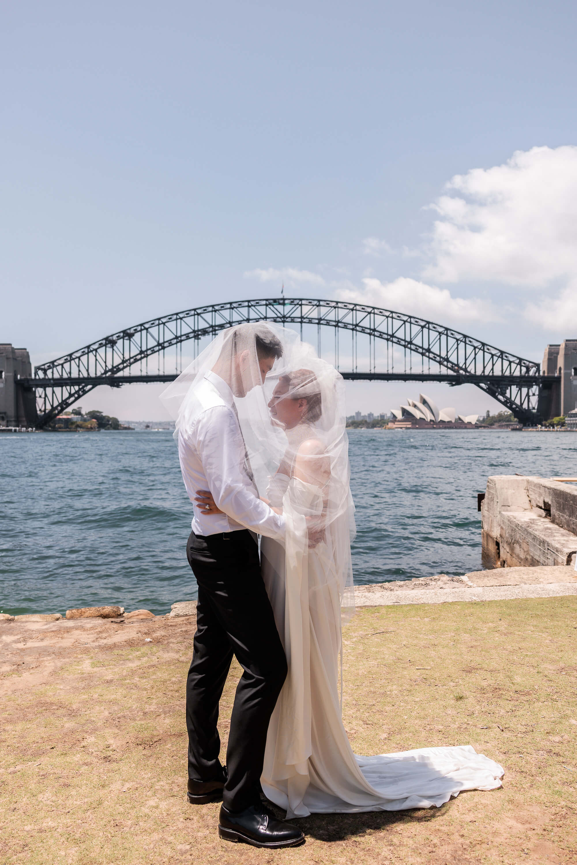 Melanie and Dai stand together beneath Melanie’s veil by the water, with Sydney Harbour Bridge and the Opera House in the background.