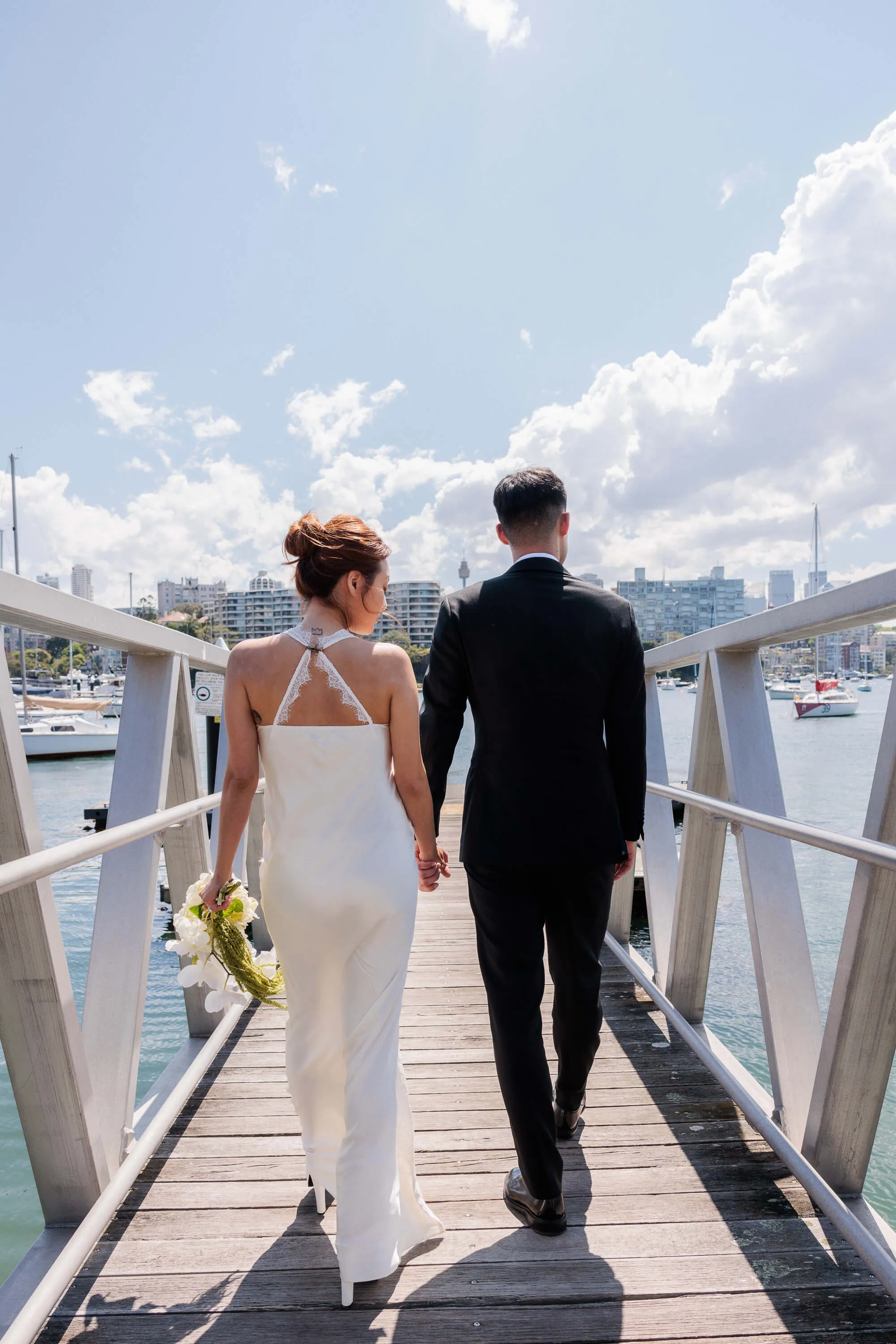 Melanie and Dai walk away together along the marina, holding hands as boats and the city skyline frame the background | EDG Studios