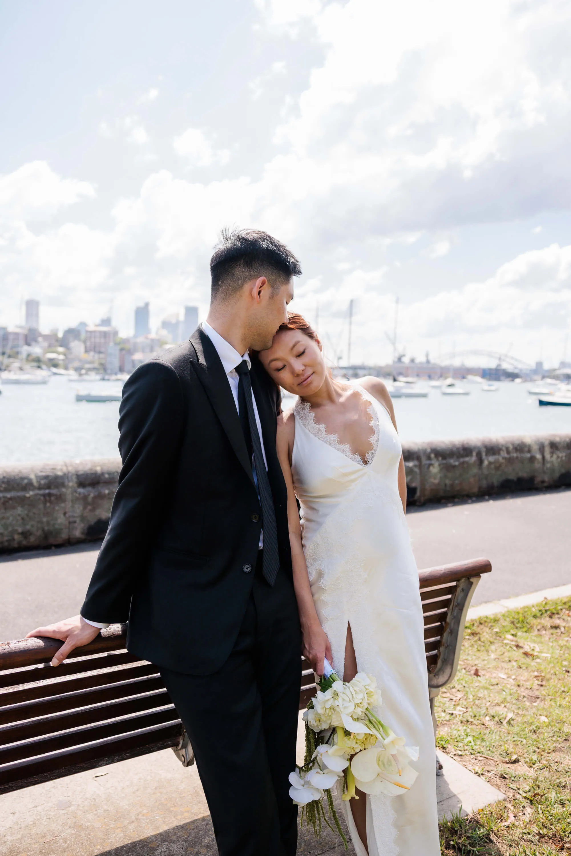 Melanie rests her head on Dai’s shoulder as they stand beside a park bench overlooking Sydney Harbour | EDG Studios