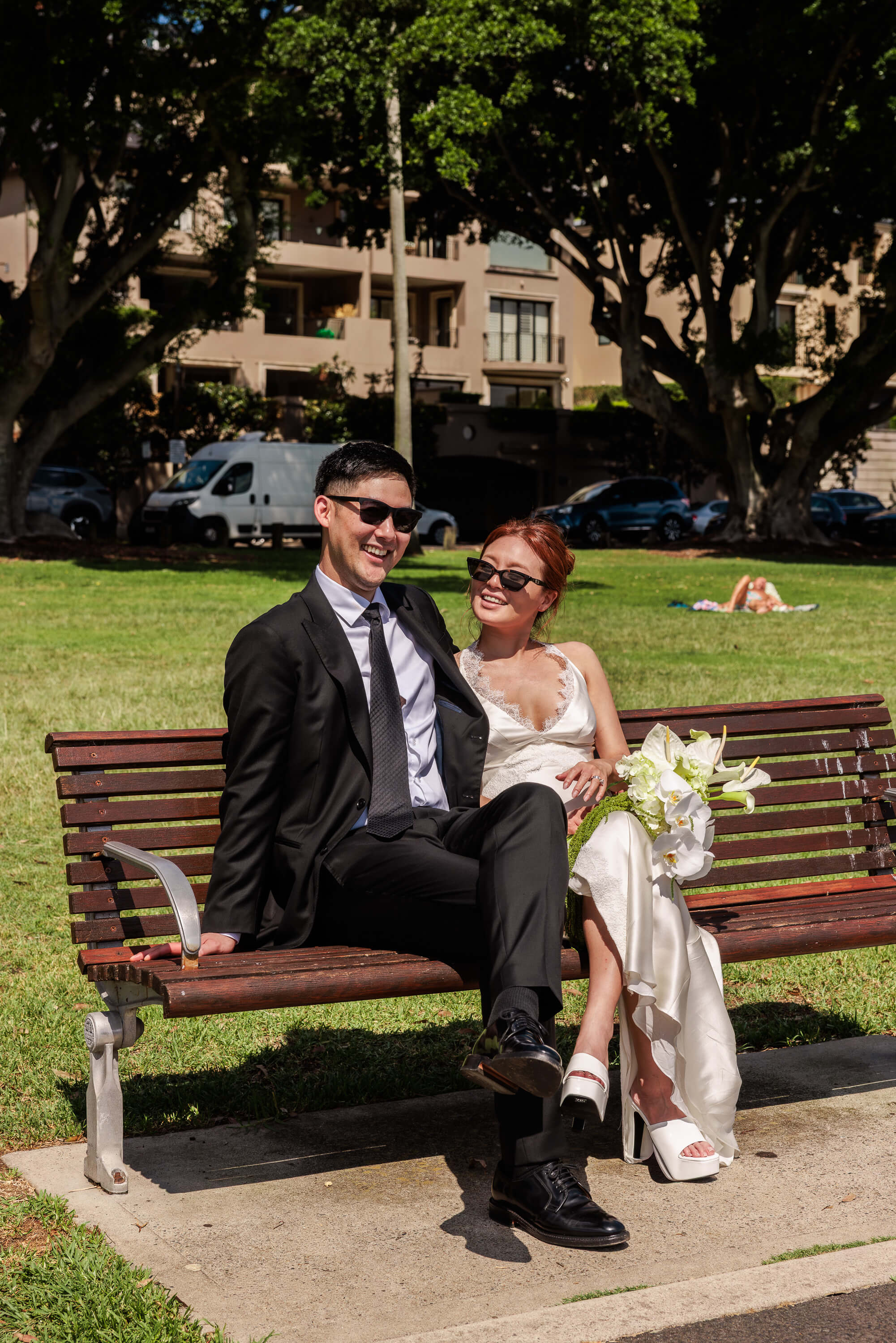 Melanie and Dai sit together on a park bench, relaxed and smiling during their wedding portraits.