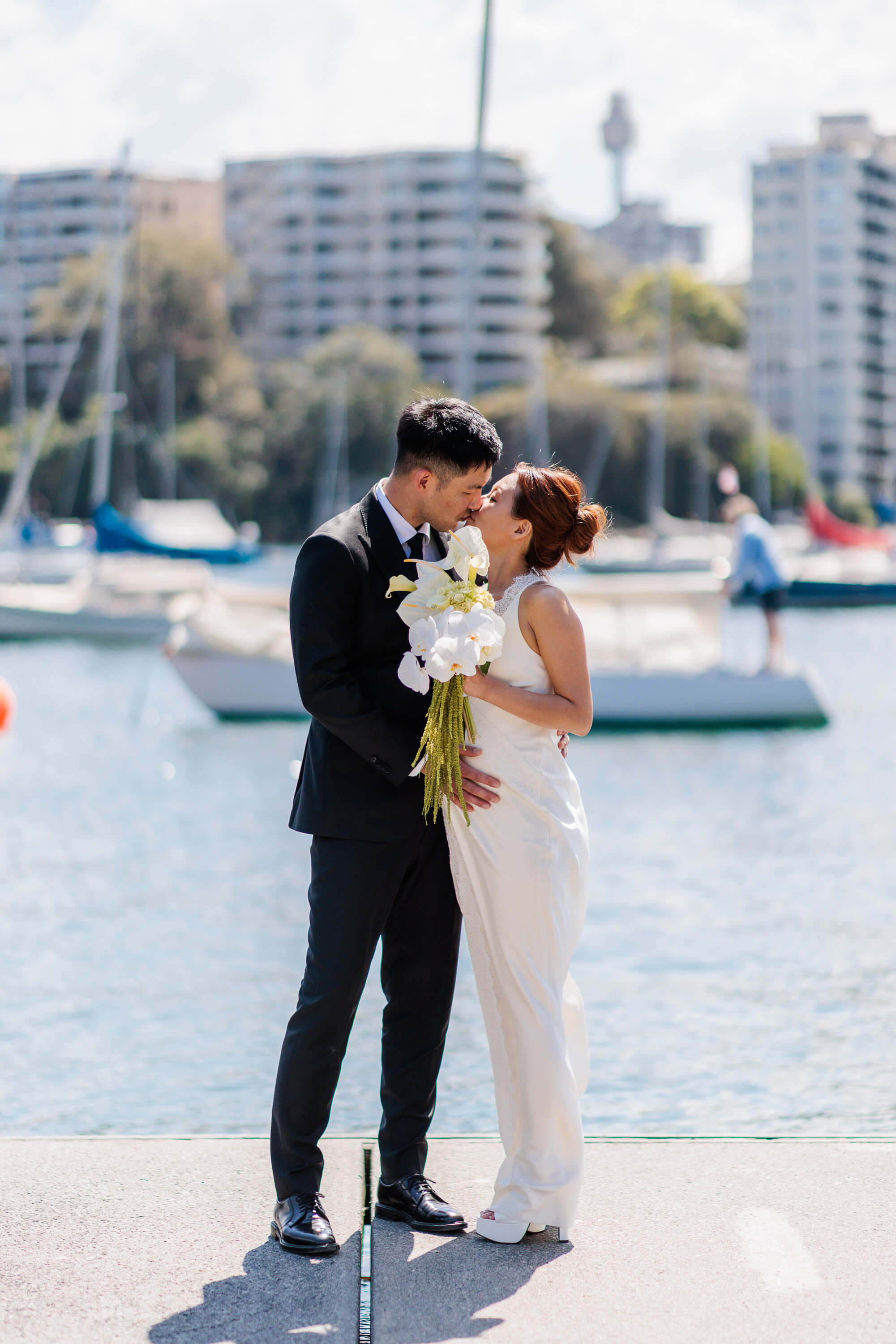 Melanie and Dai share a kiss by the water, with moored boats and the city skyline softly blurred in the background.