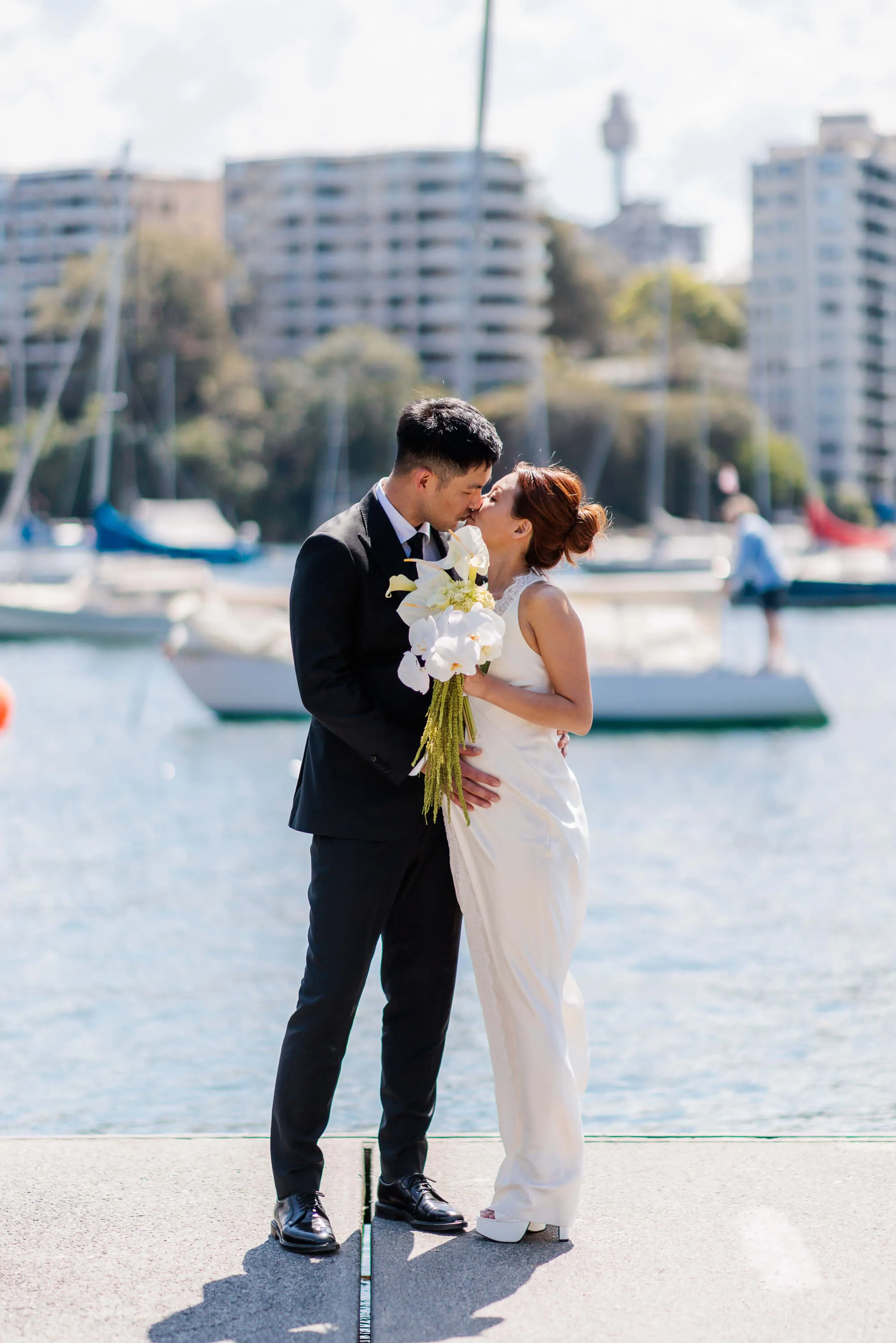 Melanie and Dai share a kiss by the water, with moored boats and the city skyline softly blurred in the background | EDG Studios