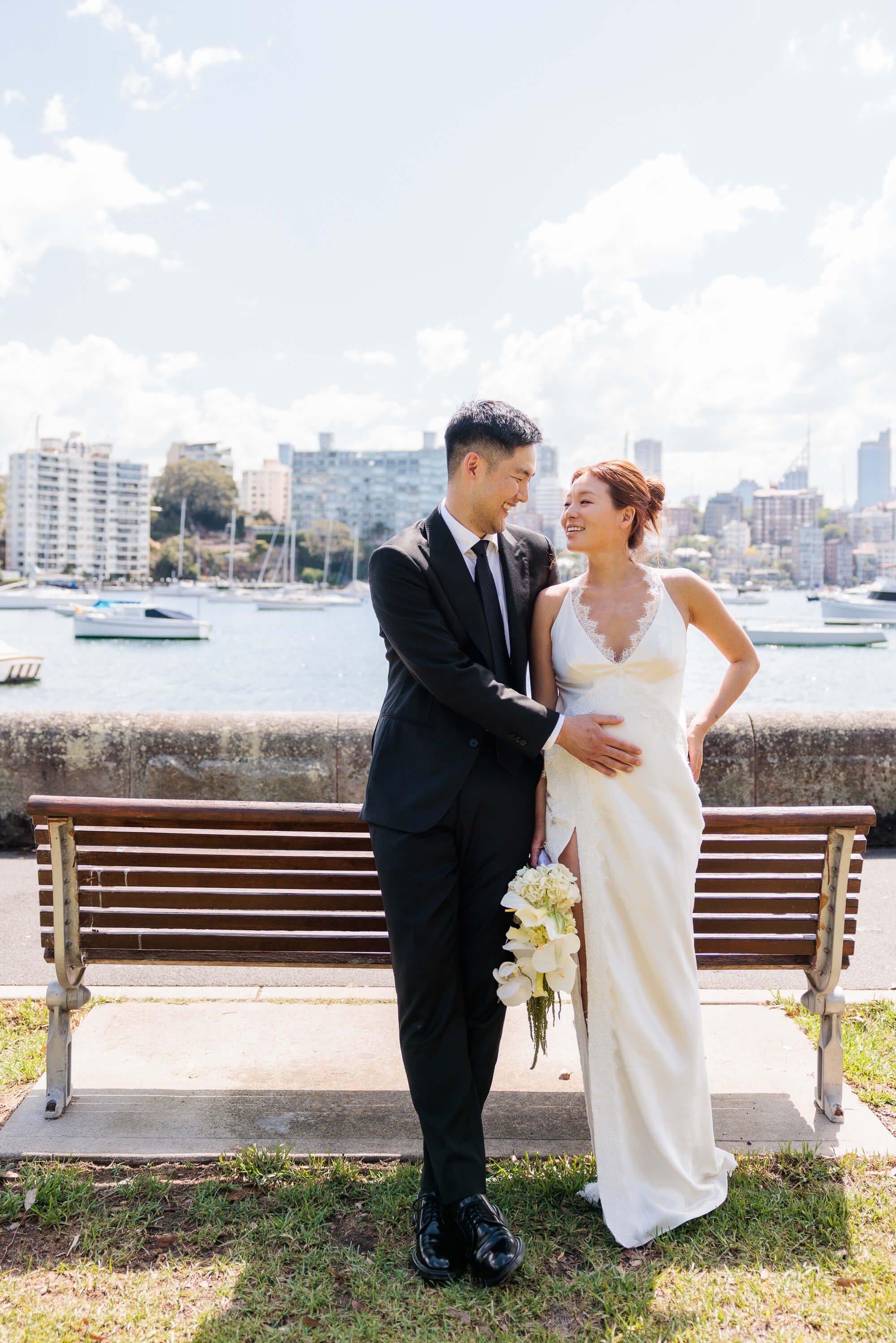 Melanie and Dai stand together by a park bench, smiling at one another during post ceremony portraits.