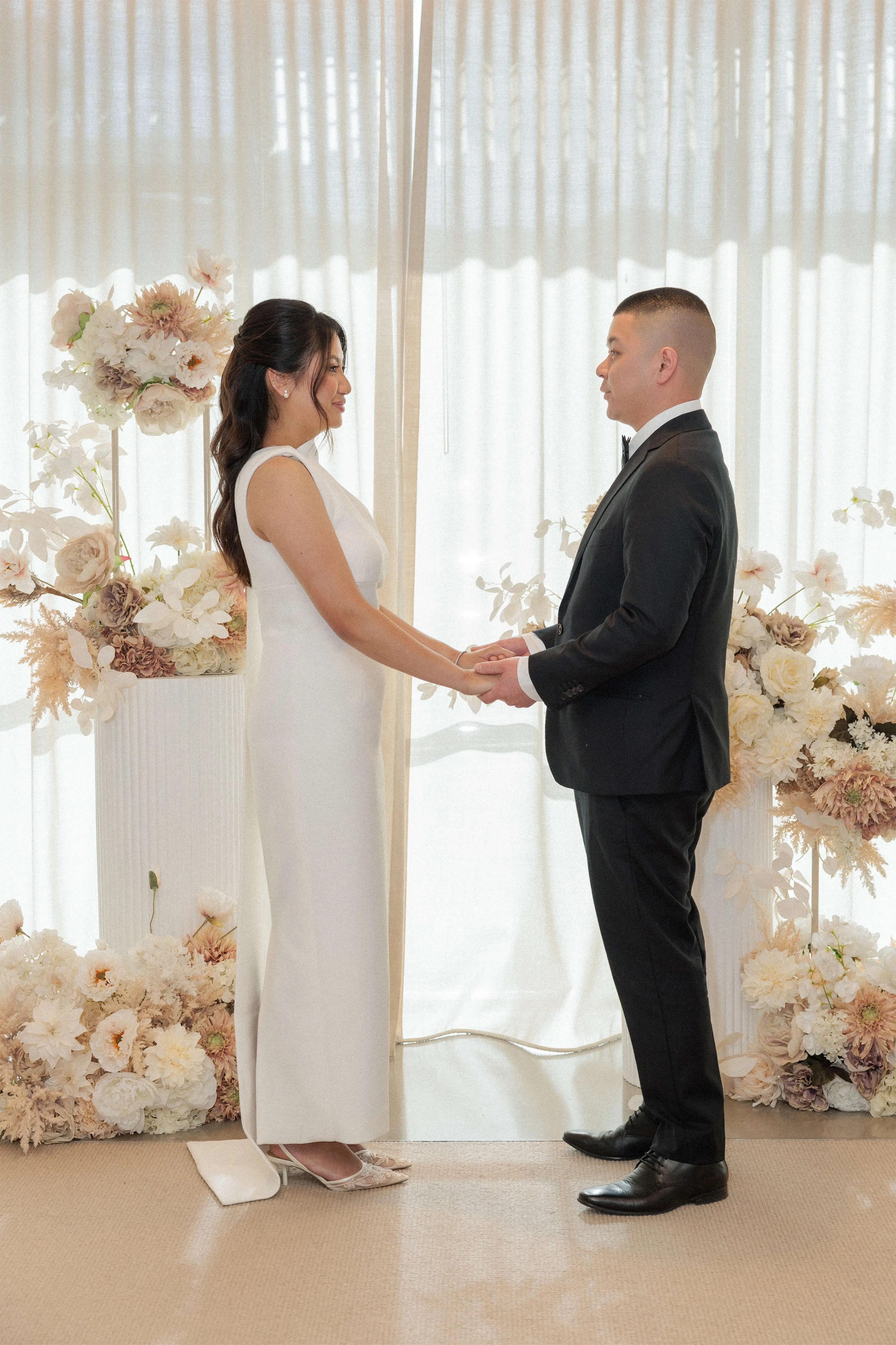 Bride and groom face each other holding hands during their indoor ceremony, framed by soft floral arrangements and sheer curtains | EDG Studios