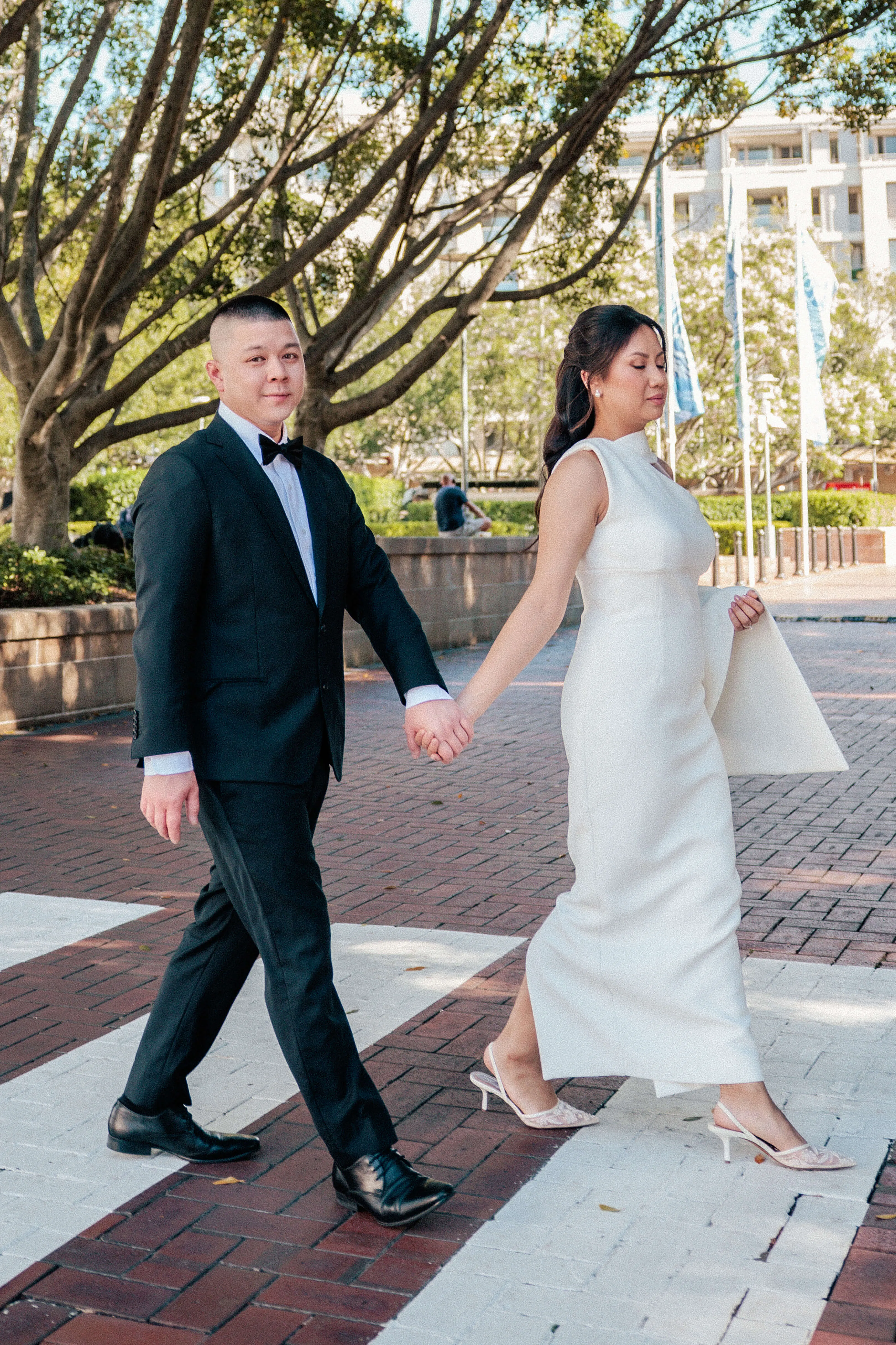 Newlyweds walk hand in hand across a red brick path, surrounded by greenery and architecture, exuding elegance and joy | EDG Studios