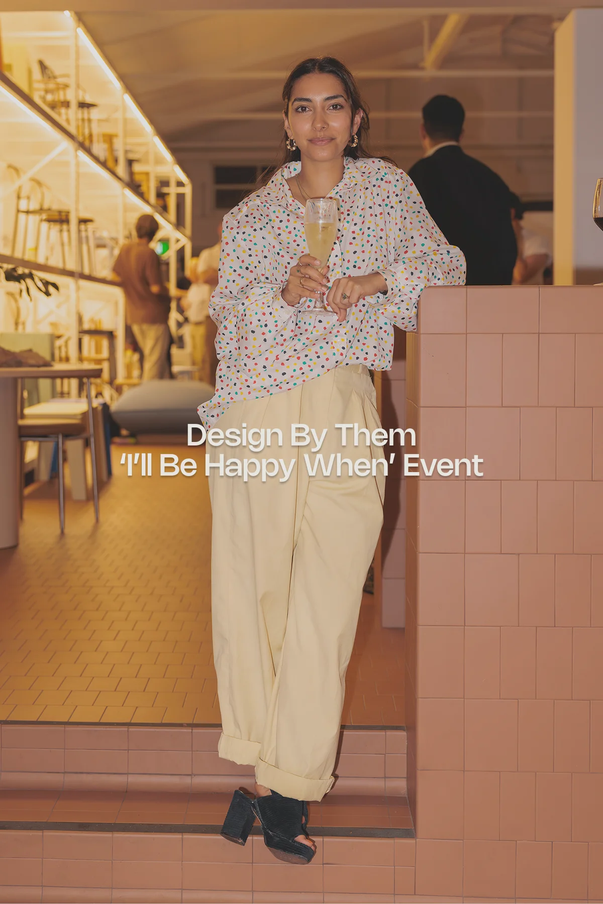 Guest attendee at the Design By Them “I’ll Be Happy When” event, held in a modern furniture showroom with designer seating, terrazzo tables, and branded signage in bold white type at the center.