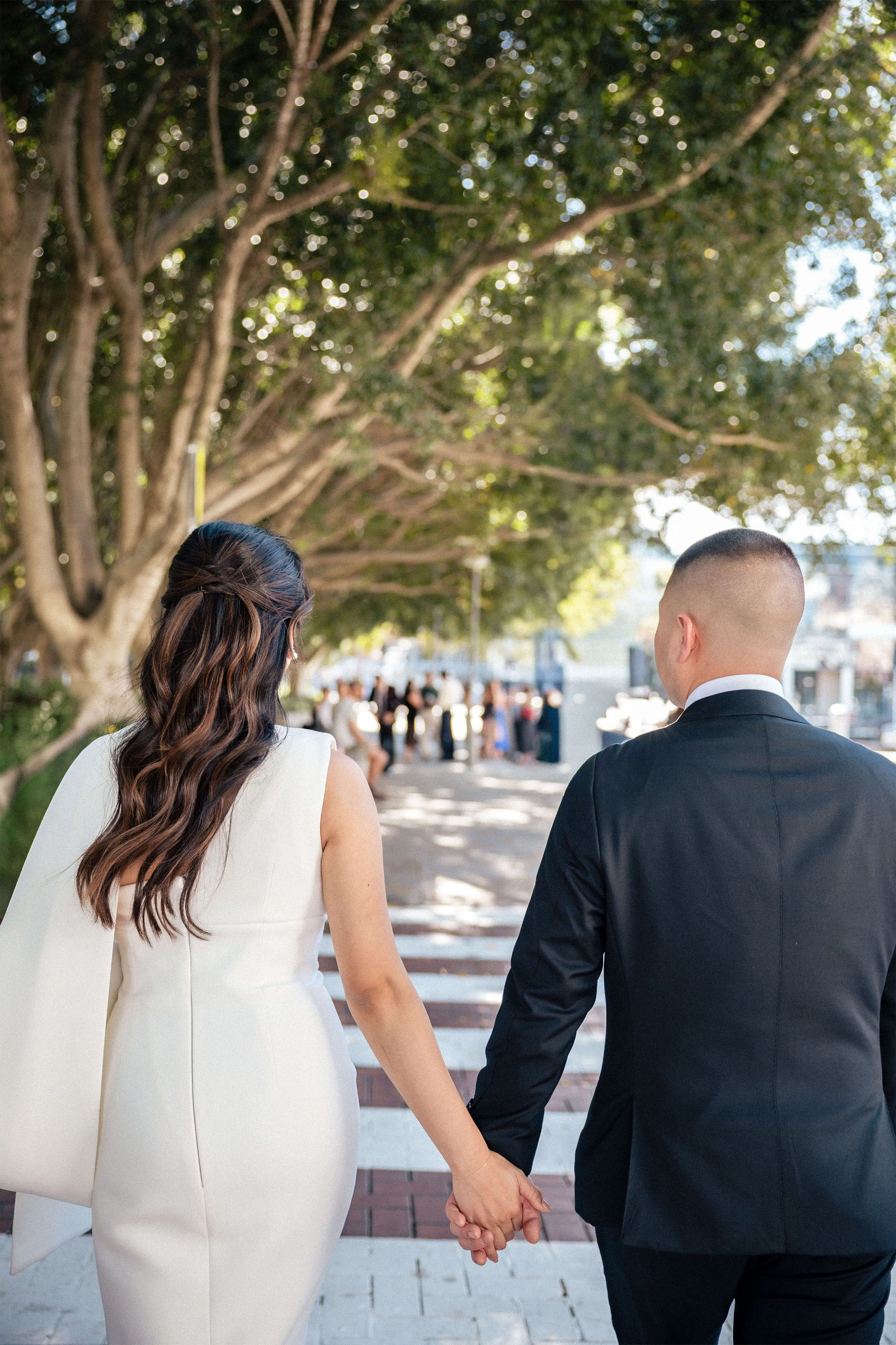 Bride and groom holding hands walking down a tree-lined street, captured from behind | EDG Studios