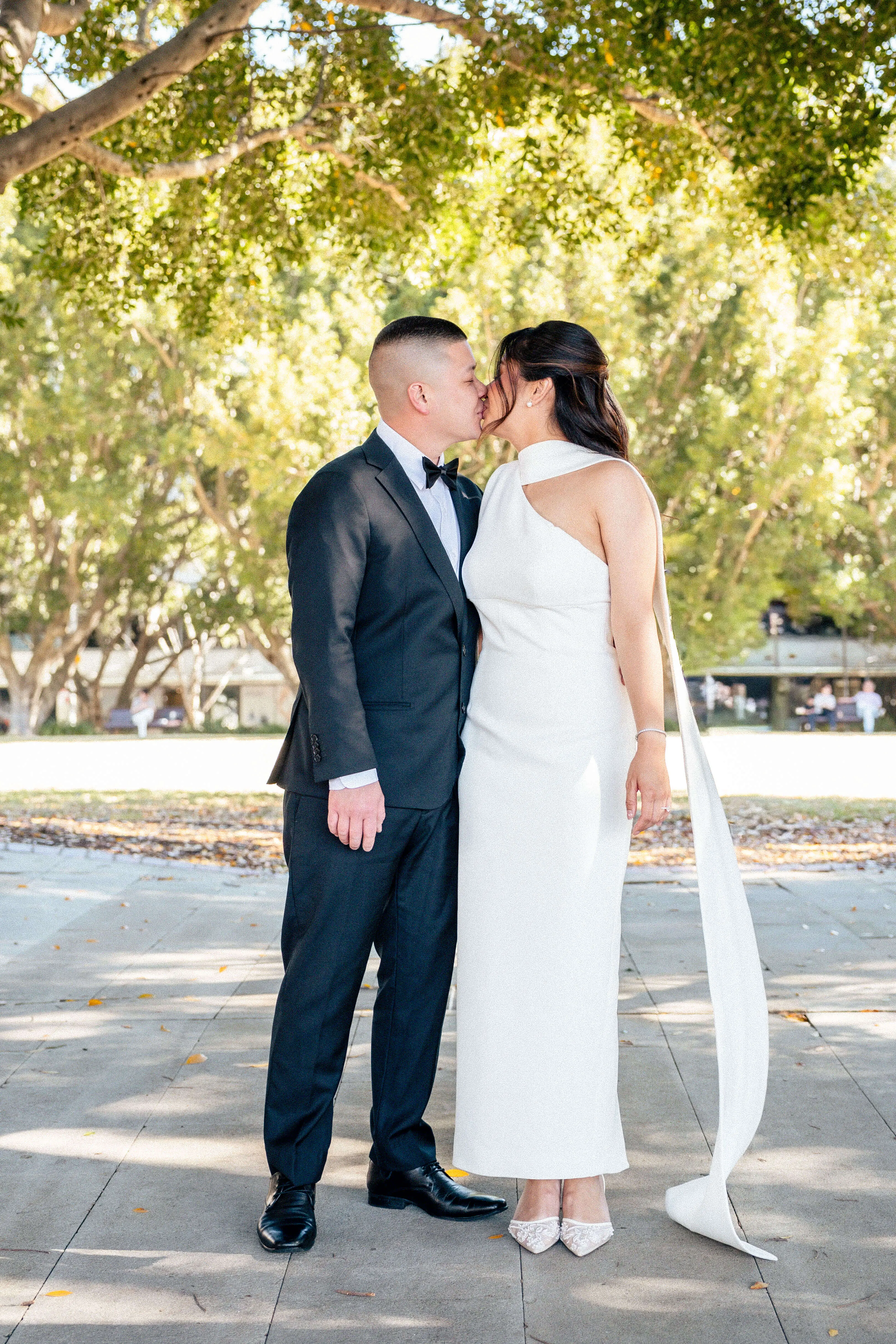Bride and groom share a kiss under a canopy of sun-drenched trees, dressed in a white modern gown and classic black tuxedo | EDG Studios