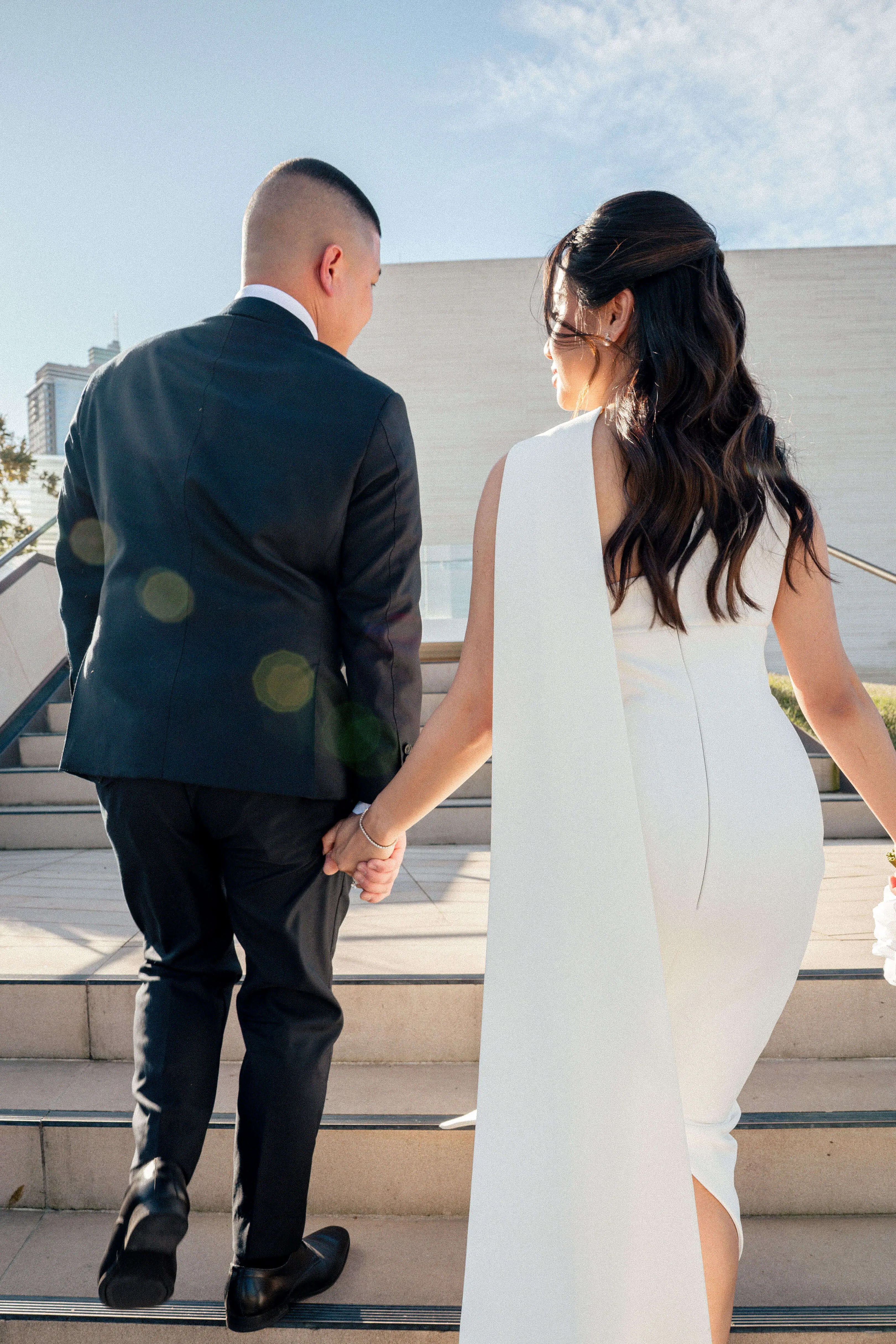 Candid shot of the couple walking up stairs, with lens flare and the bride glancing back over her shoulder, dress detail trailing behind | EDG Studios