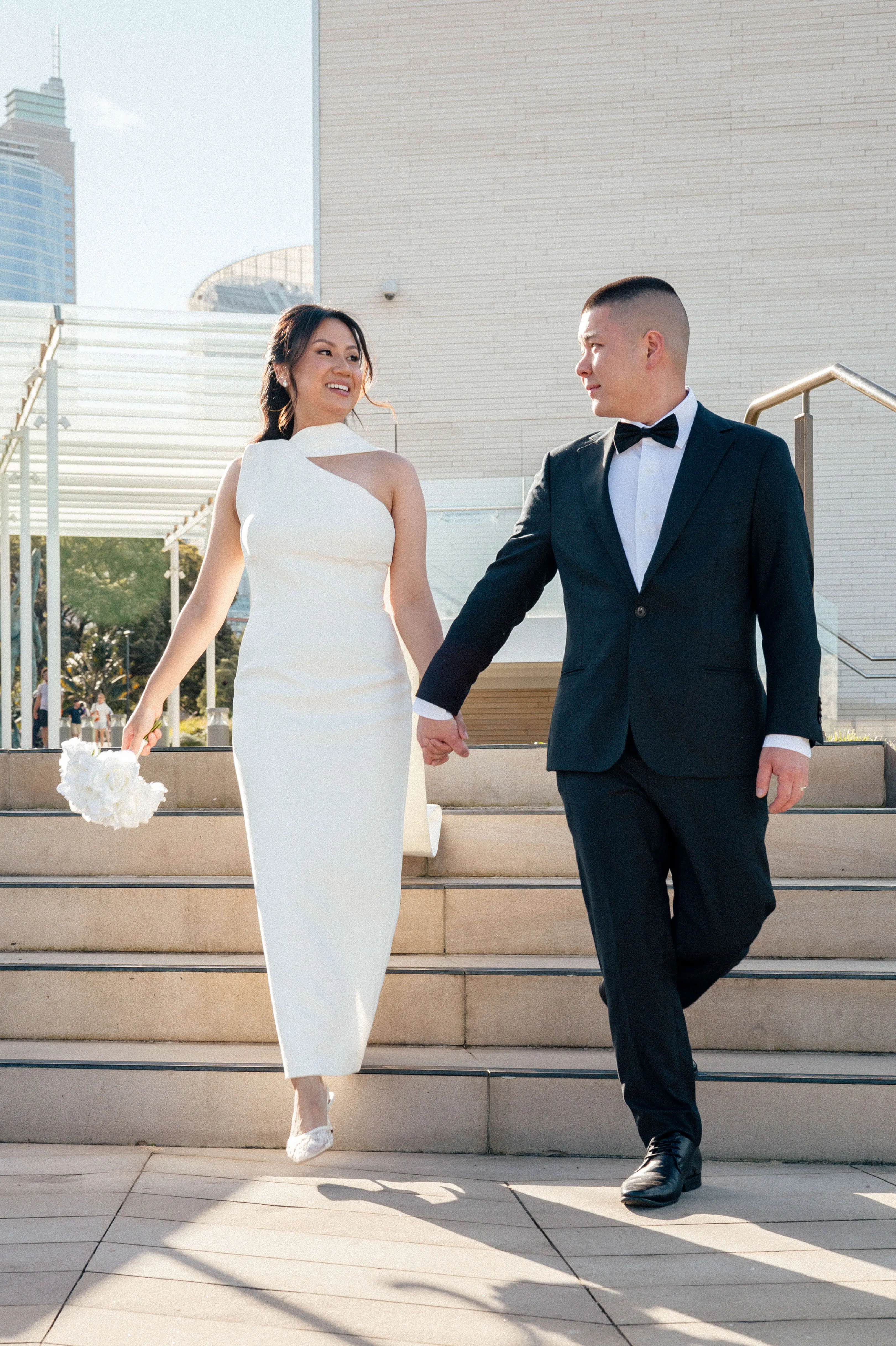 Bride and groom walk down sunlit stone steps, smiling at each other, with a bouquet in hand and a modern cityscape in the background | EDG Studios