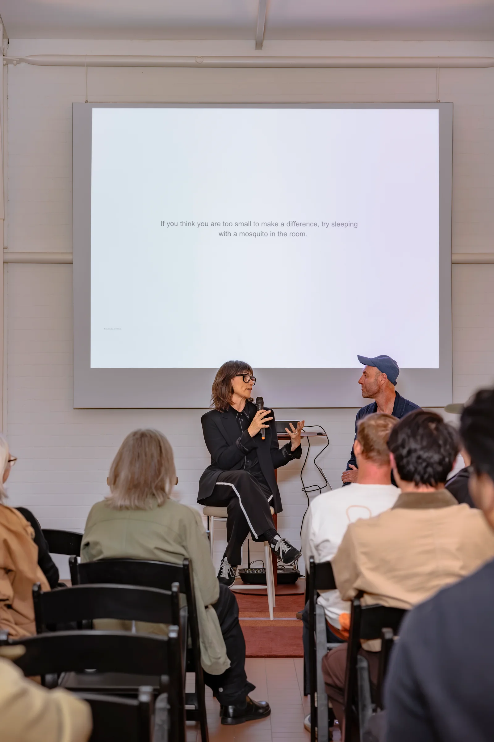 Two speakers in conversation beneath the projected presentation at Design By Them, photographed by EDG Studios.