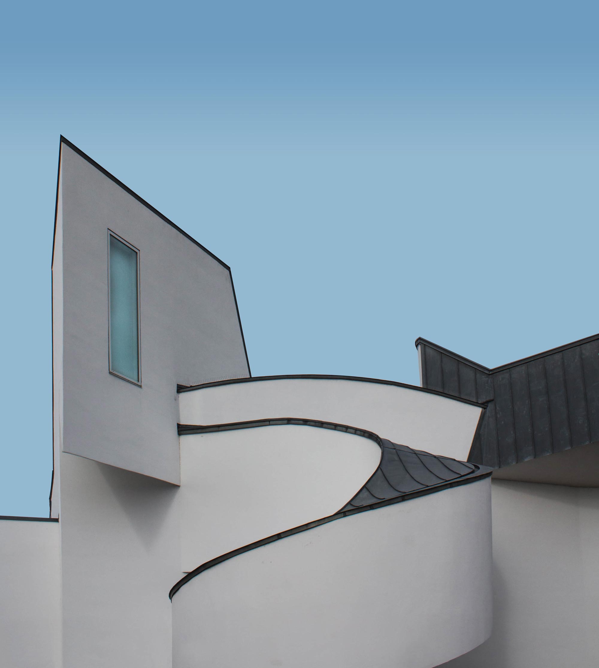 Modern white building with curved balcony and a single narrow window against a clear blue sky.