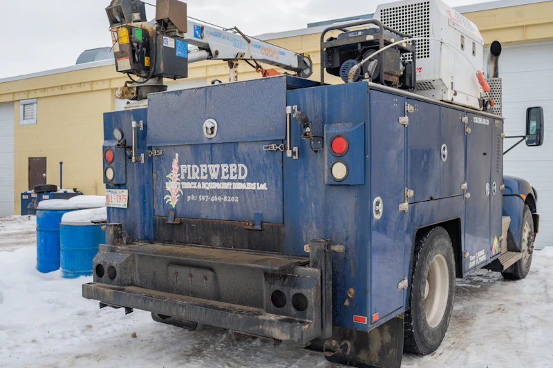 A blue utility truck labeled "FIREWEED Truck & Equipment Repairs Ltd." is parked on a snowy lot beside blue barrels and a yellow building, with visible equipment including a Cobra crane and a large white generator on top.