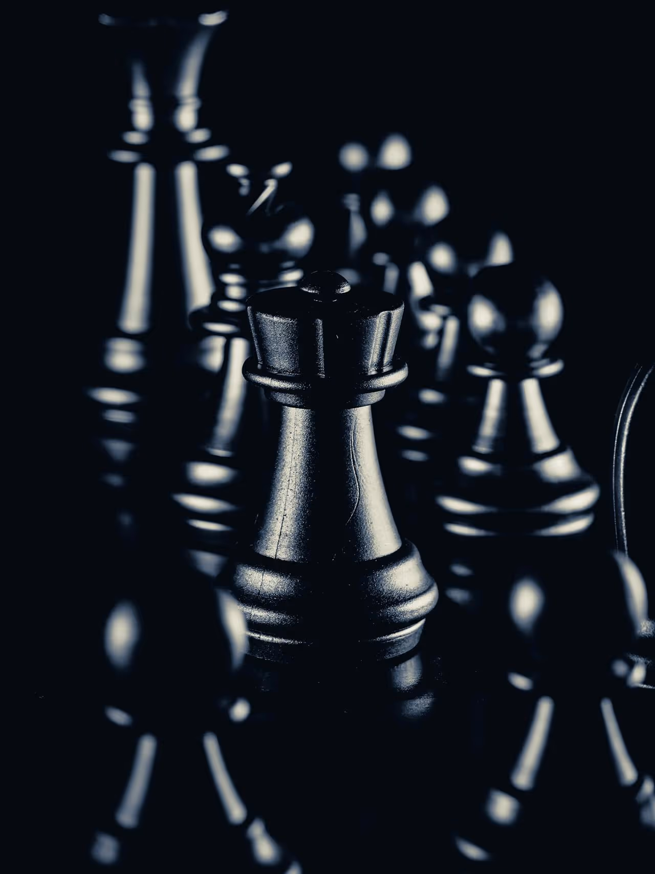 A close-up, dramatic image of black chess pieces, with the queen prominently in the center, surrounded by other pieces, against a dark background.