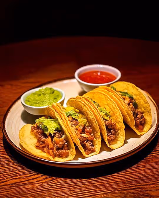 Four tacos on a plate with a side of creamy guacamole and salsa, inviting you to dig in and enjoy.