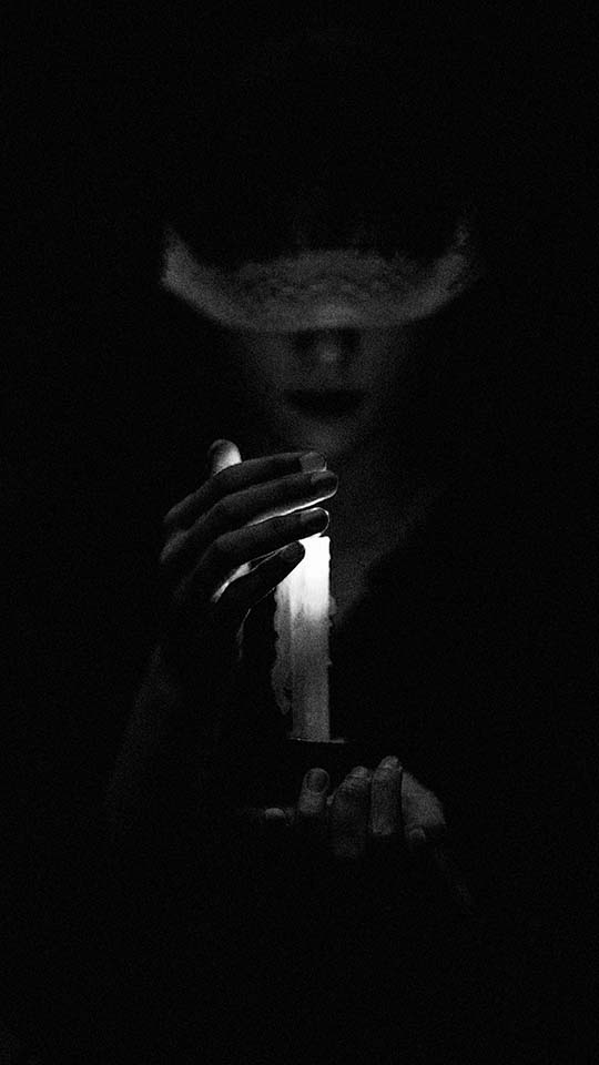 A blindfolded woman in shadow holds a lit candle, with their face partially obscured and dimly illuminated by the candlelight in the darkness. The mood is mysterious and somber.