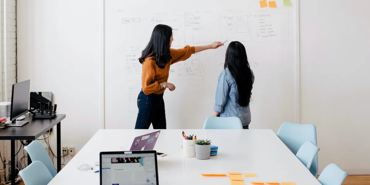 Two people stand before a whiteboard with sketches of a website design.
