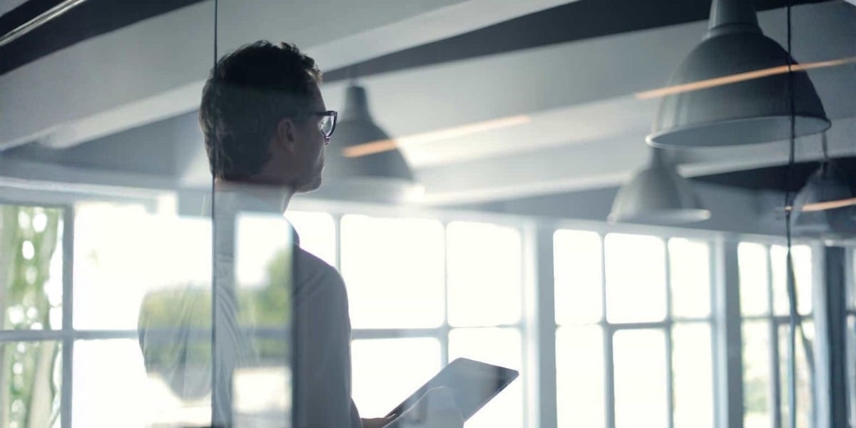 A man in glasses holds a tablet while standing in an office.