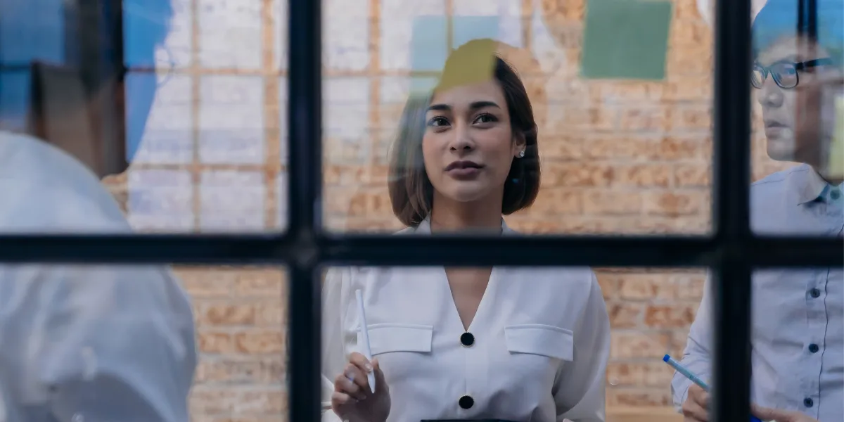 Woman in a white shirt gestures with a pen through a window.
