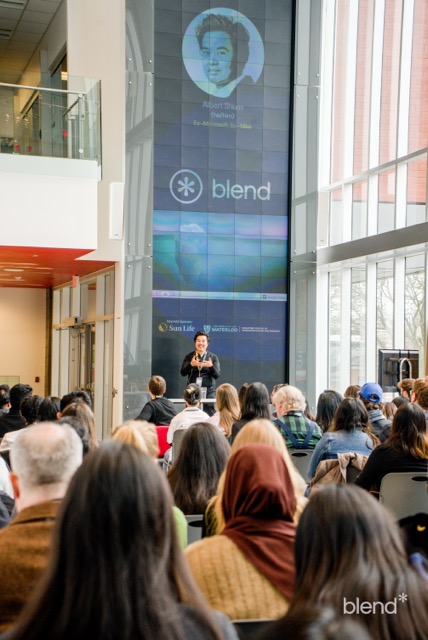 The keynote speaker stands on stage  in the Atrium of the Stratford School.  The crowd sits in front, listening to their presentation.