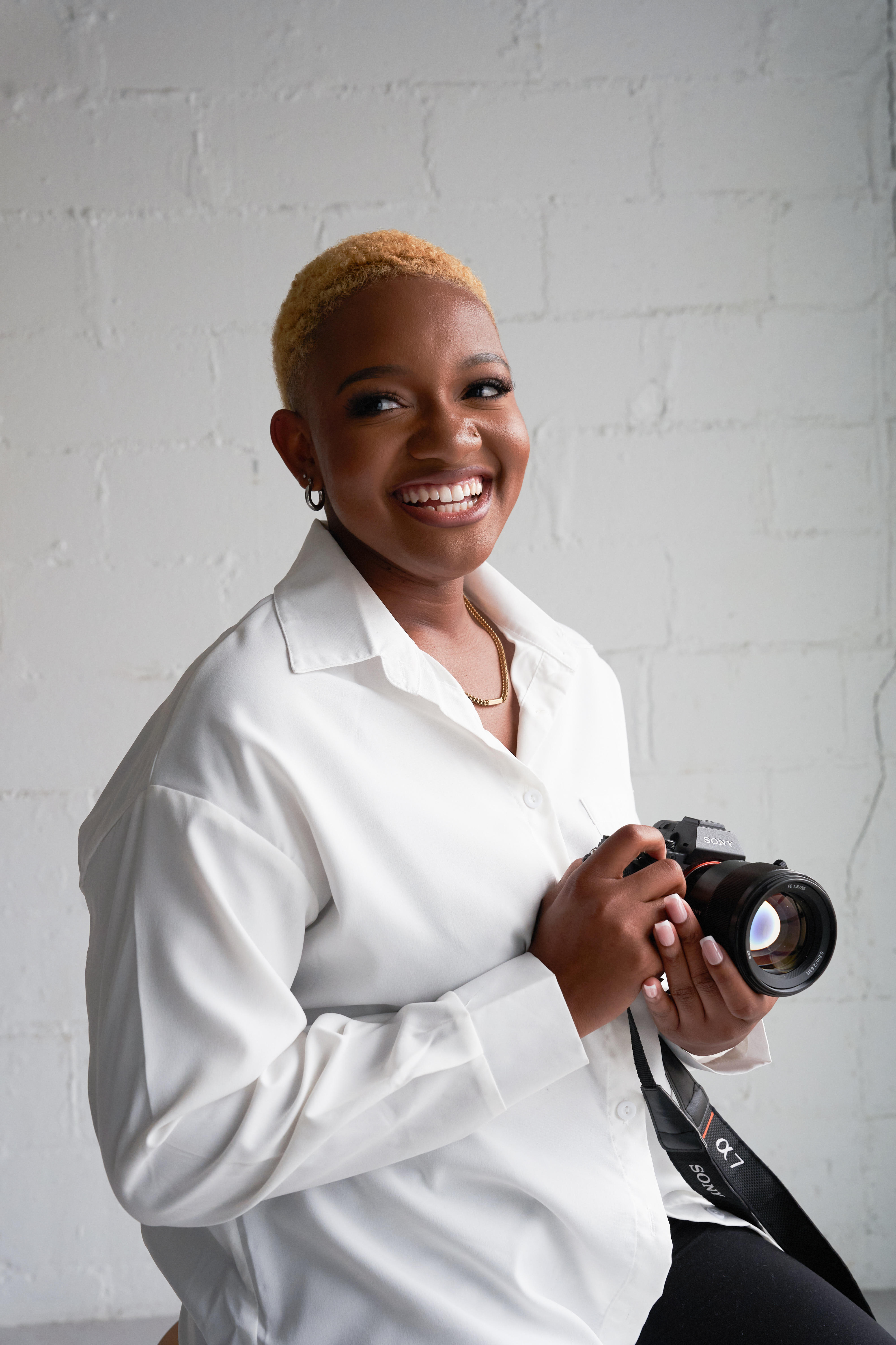 Kalena, a black woman with short hair wearing a white button down shirt, smiles at the camera. She is holding a camera in her hands.