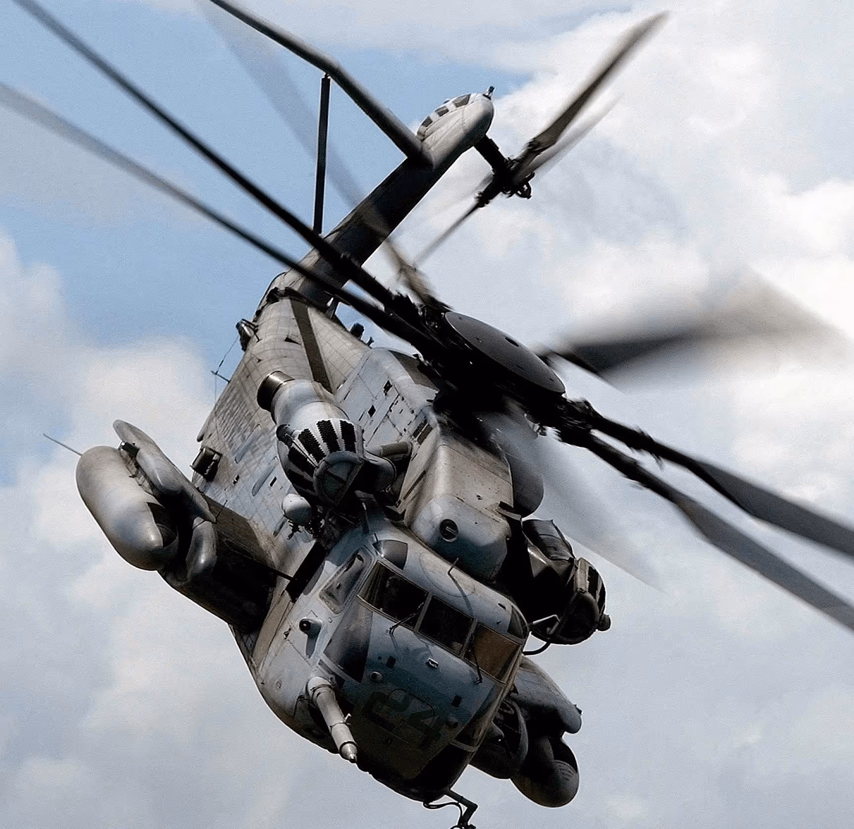 Military helicopter in flight viewed from front below with spinning rotor blades against a cloudy sky.
