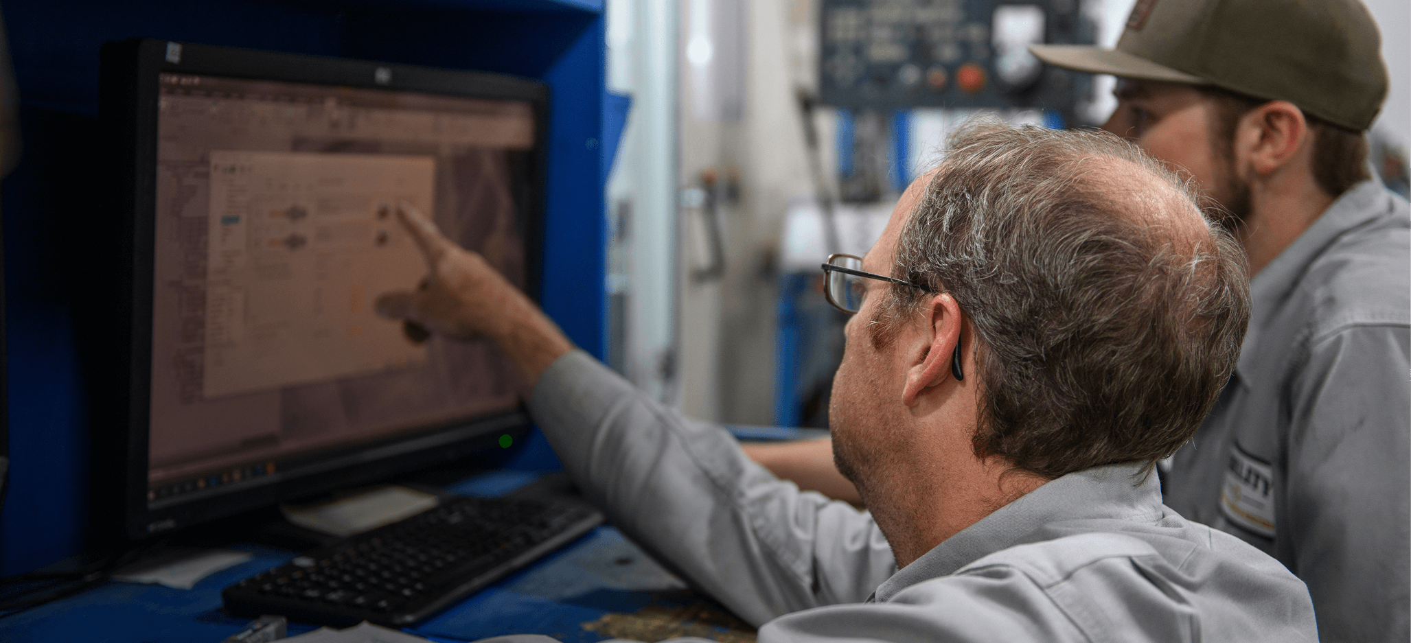 Two technicians in work uniforms examining and discussing information on a computer screen in an industrial setting.