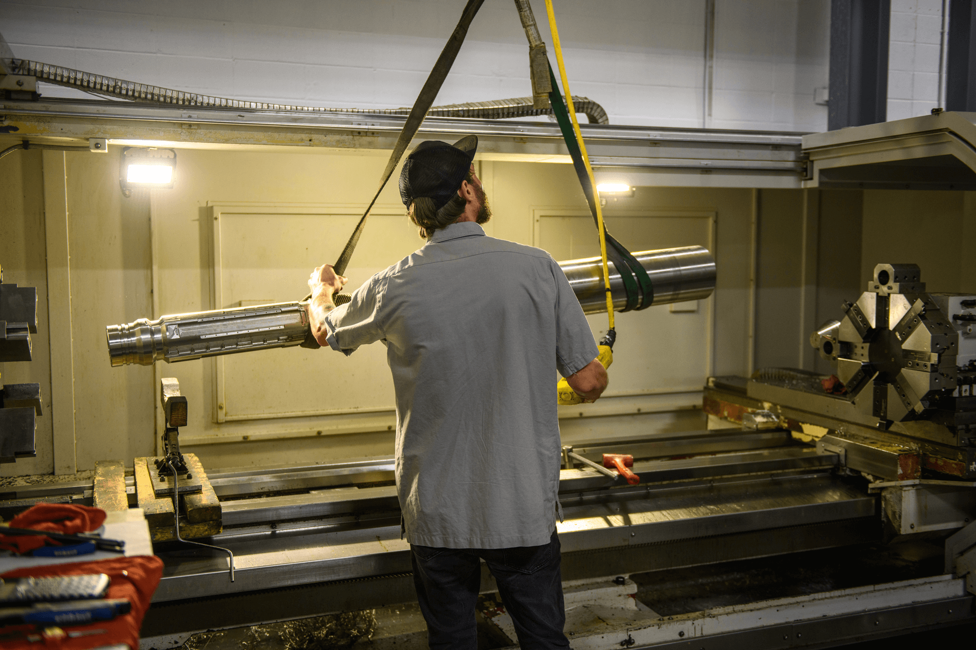 Worker wearing a cap and gray shirt guiding a large metal cylindrical part suspended by straps inside a factory machine.