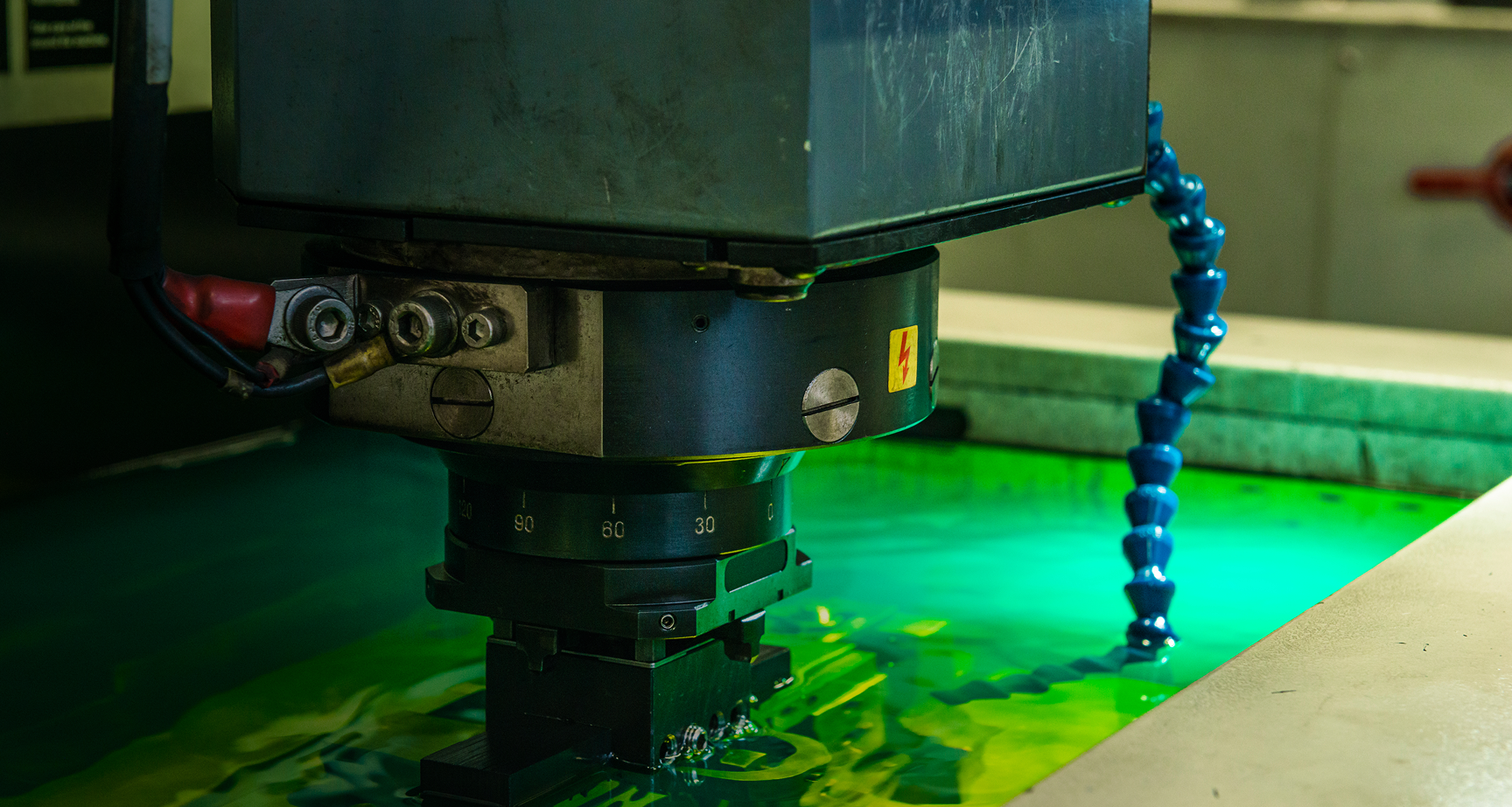 Close-up of an industrial machine part with metal connectors and a flexible blue tube over a green liquid surface.