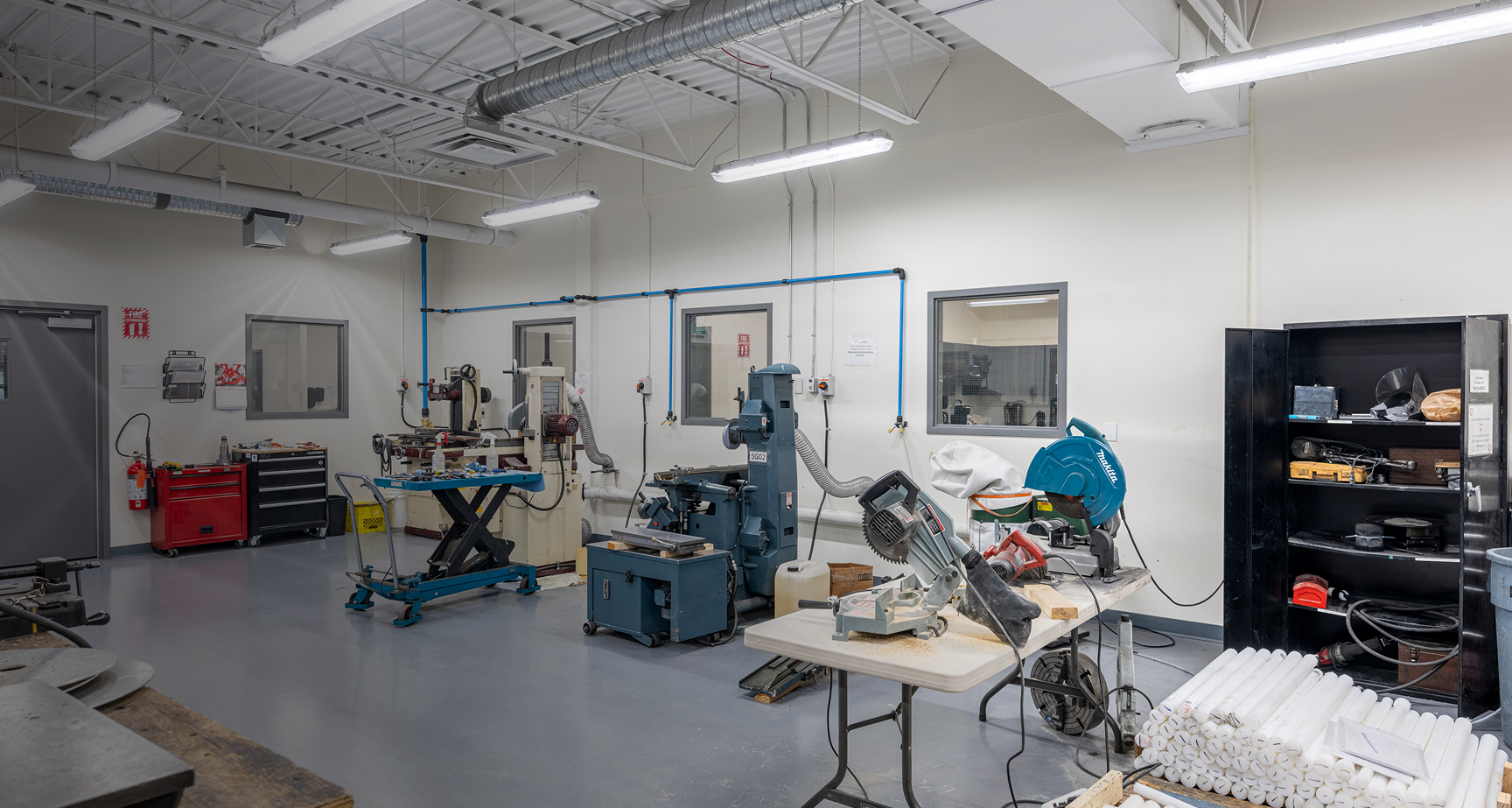 Workshop with various industrial machines, including a blue Makita chop saw on a table, tool cabinets, and shelving with equipment.