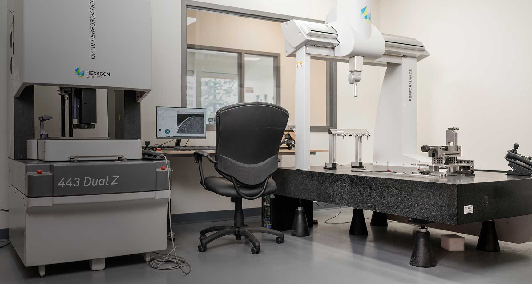 Industrial inspection room with a Hexagon Metrology machine labeled 443 Dual Z, a black office chair, a computer monitor showing microscopic images, and a large granite surface plate with mechanical fixtures.