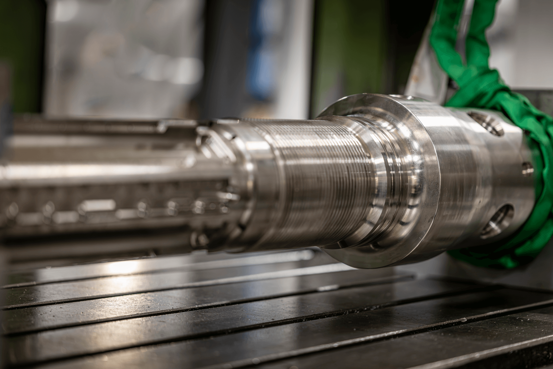 Close-up of a shiny metal industrial machine part with threaded sections and green lifting straps in a workshop.