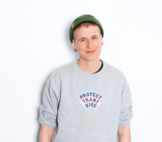 Freddie* trägt eine grüne Mütze und einen grauen Pulli mit der Aufschrift „Protect trans kids“